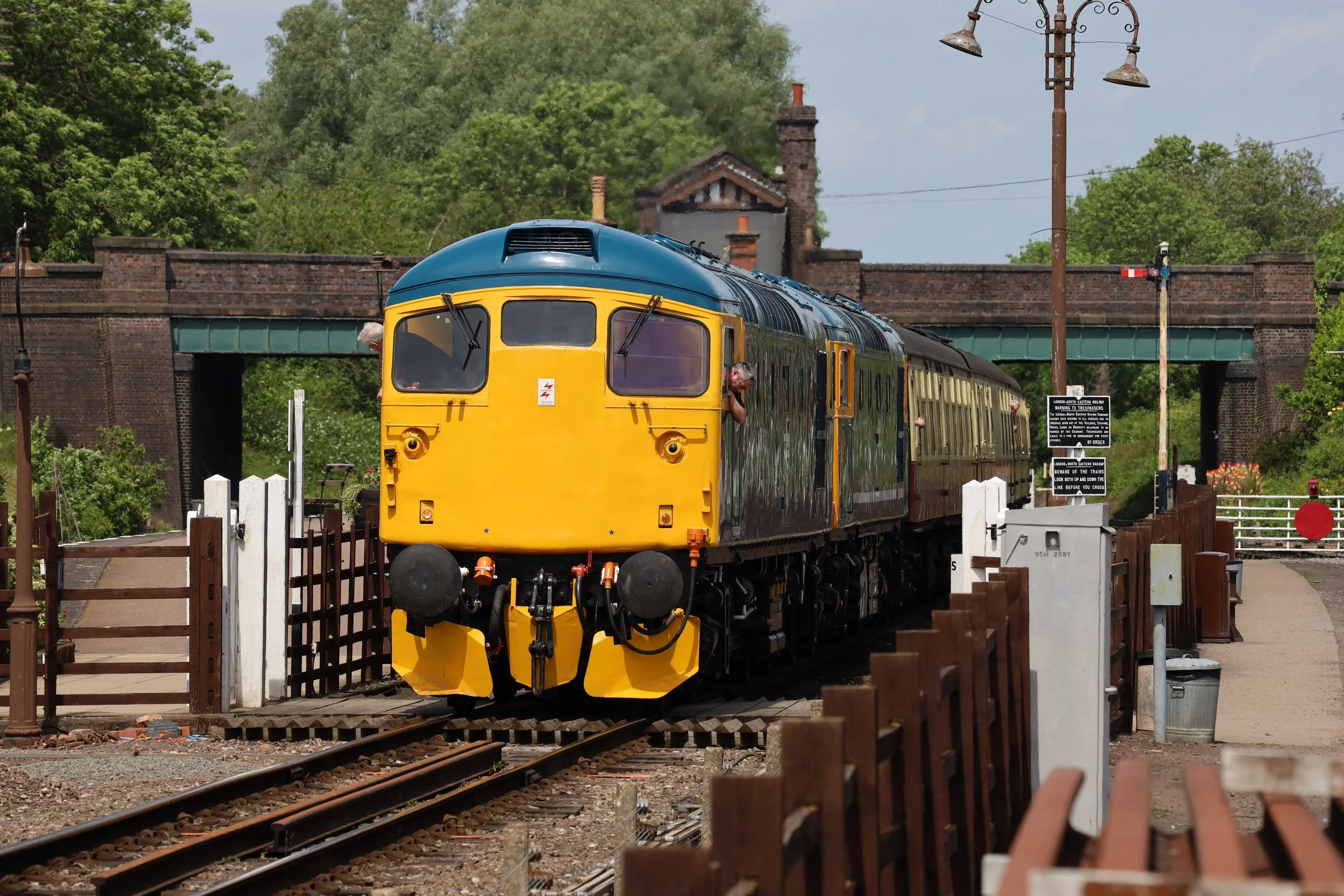 A vintage yellow and blue train is traveling on railway tracks near a rural station, with green trees and a brick bridge in the background, and various warning signs around the tracks.