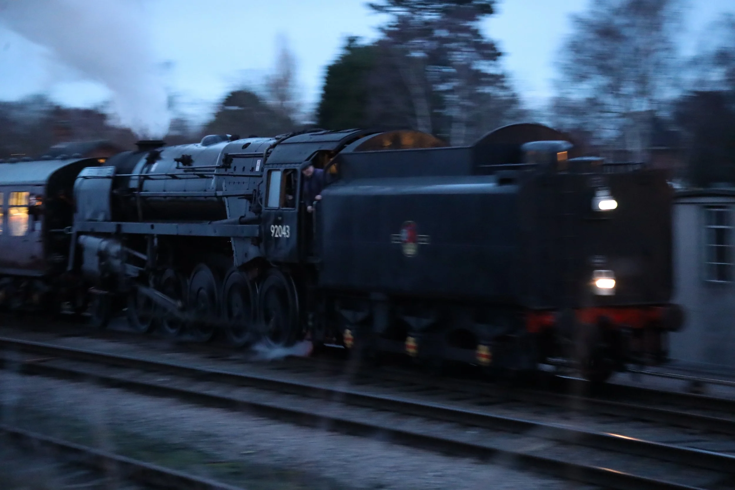 A steam locomotive train moving on railway tracks in a rural area during dusk or dawn with trees in the background.