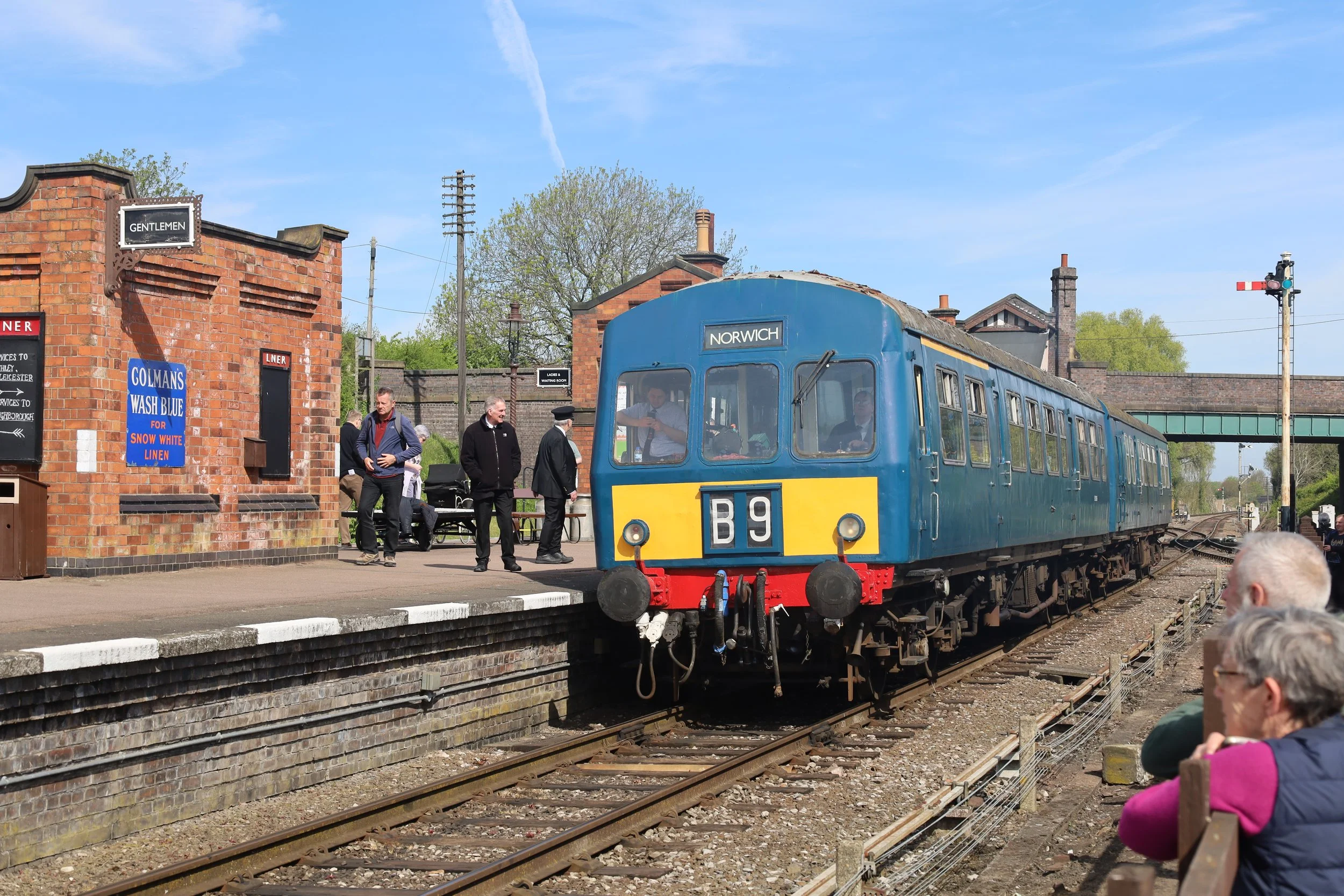 People at a train station with a vintage blue train labeled "Norwich" on the track, brick station building with signs, and a crossing signal in the background.