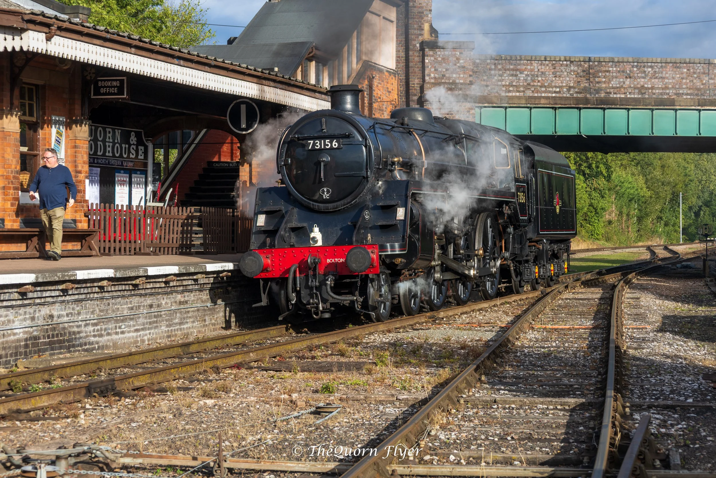 A black steam locomotive number 73156 at a train station, emitting steam, with a brick station building and a person walking on the platform.