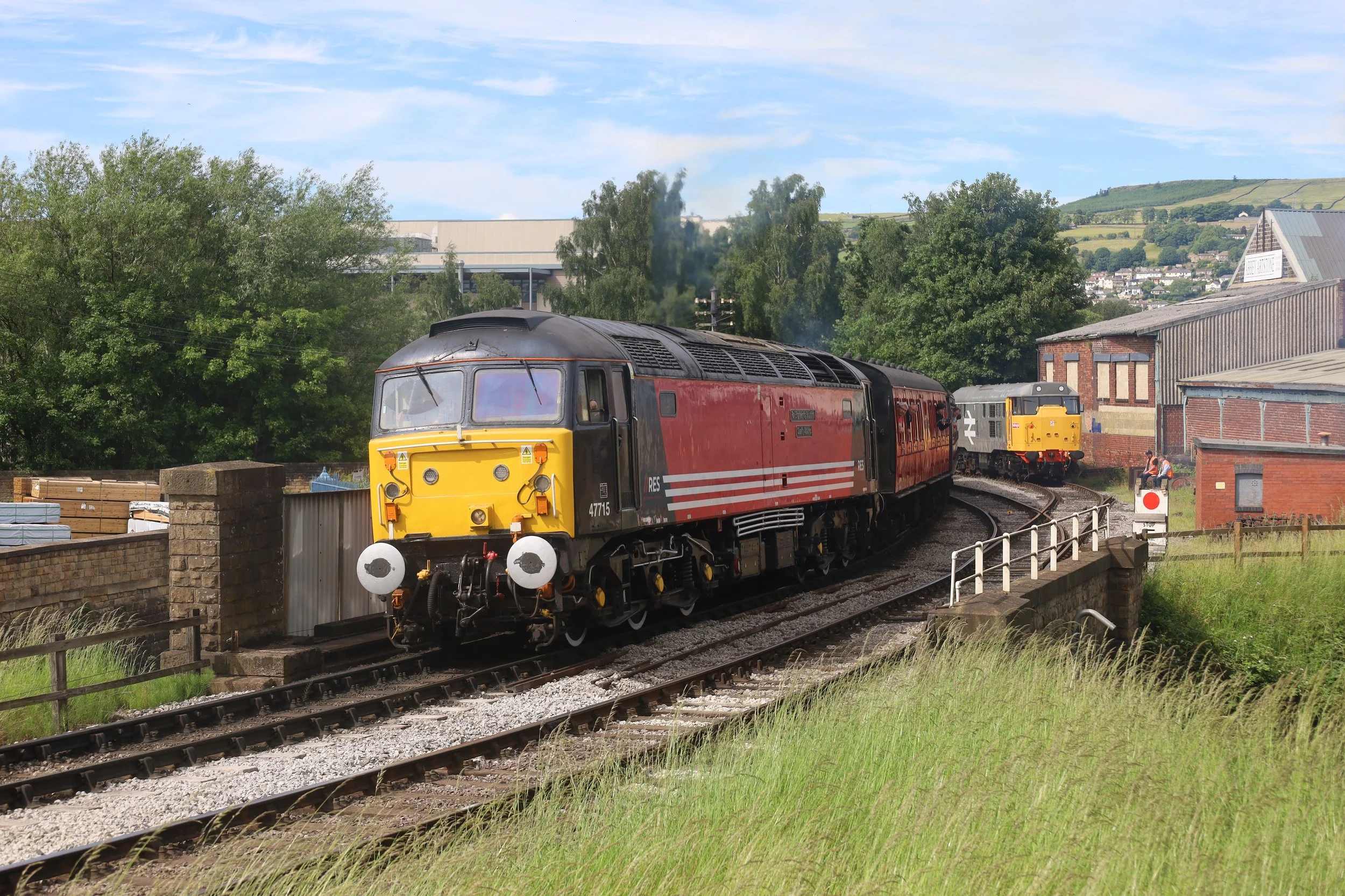 A train with a yellow front and red body traveling on a curved railway track in a rural area with green trees, buildings, and hills in the background.