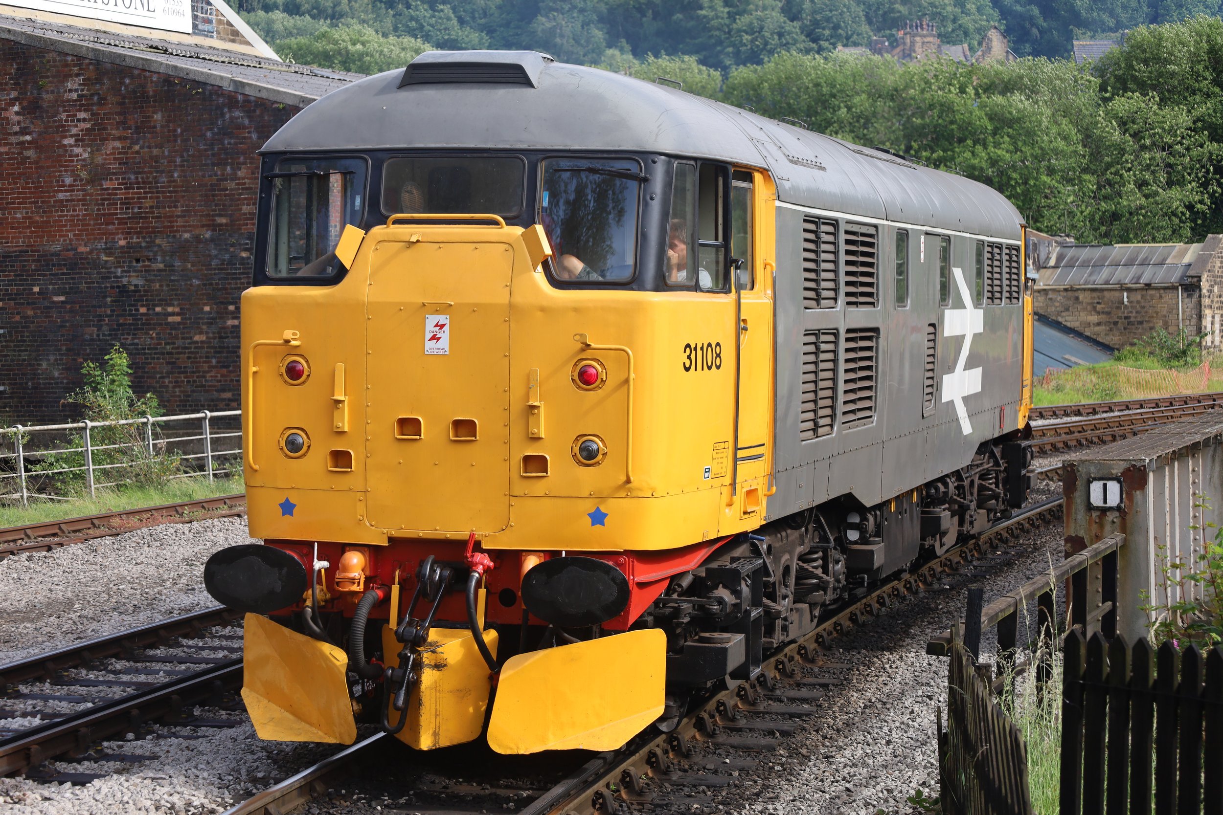 A yellow and gray train on a track with green trees and a brick building in the background.