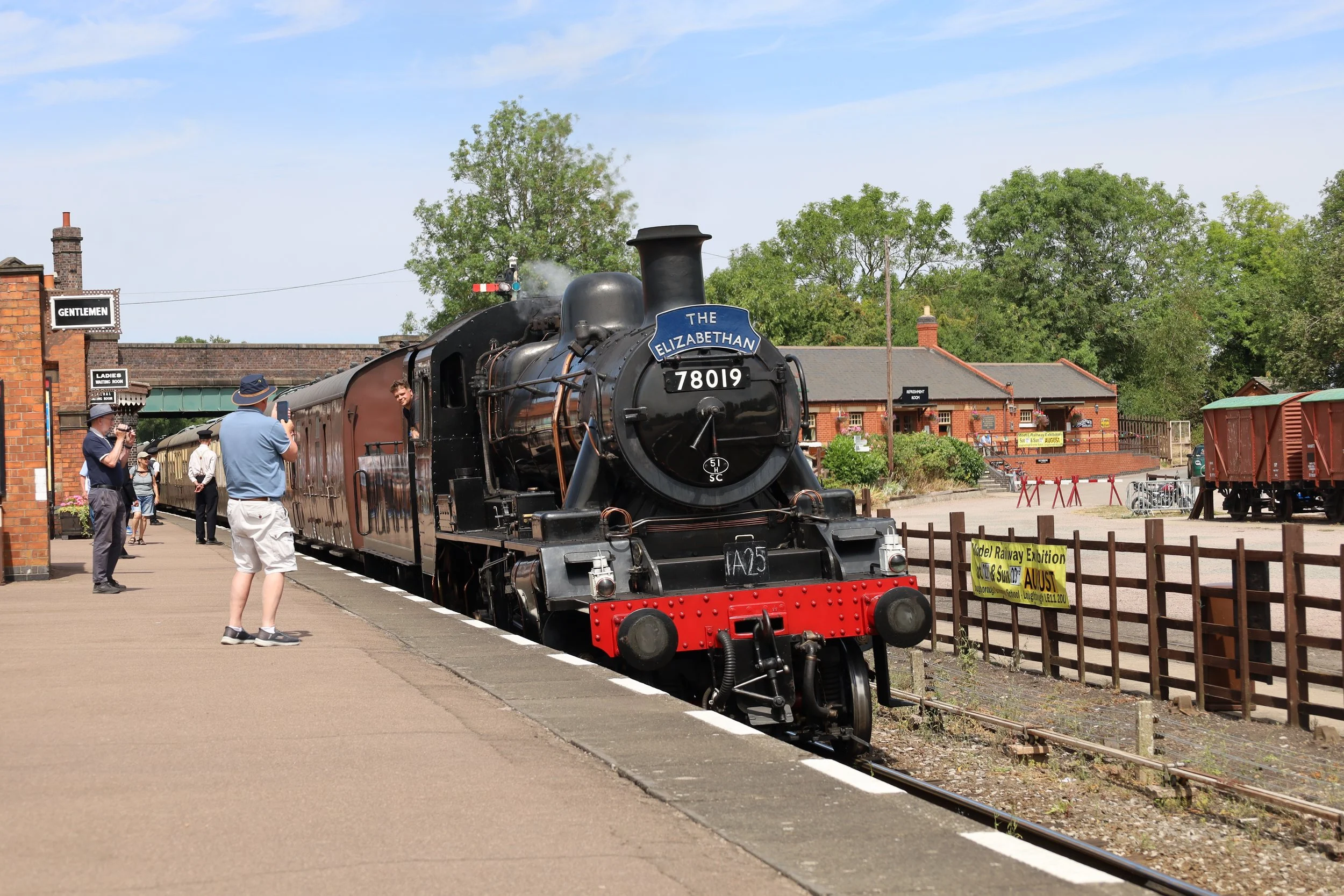 Vintage steam train named 'The Elizabethan' at a train station with people taking photos, brick station buildings, trees in the background, and signage indicating separate areas for gentlemen and ladies.