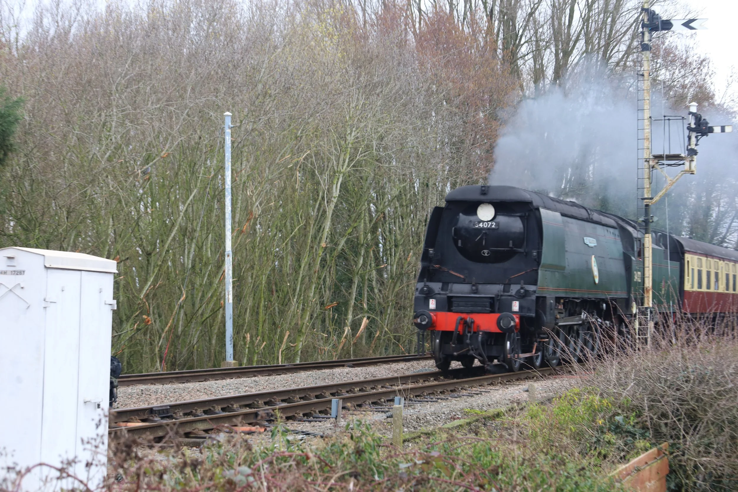 A black steam locomotive with a red buffer beam pulling a passenger carriage along railway tracks nearby a wooded area with leafless trees and railway signaling equipment.