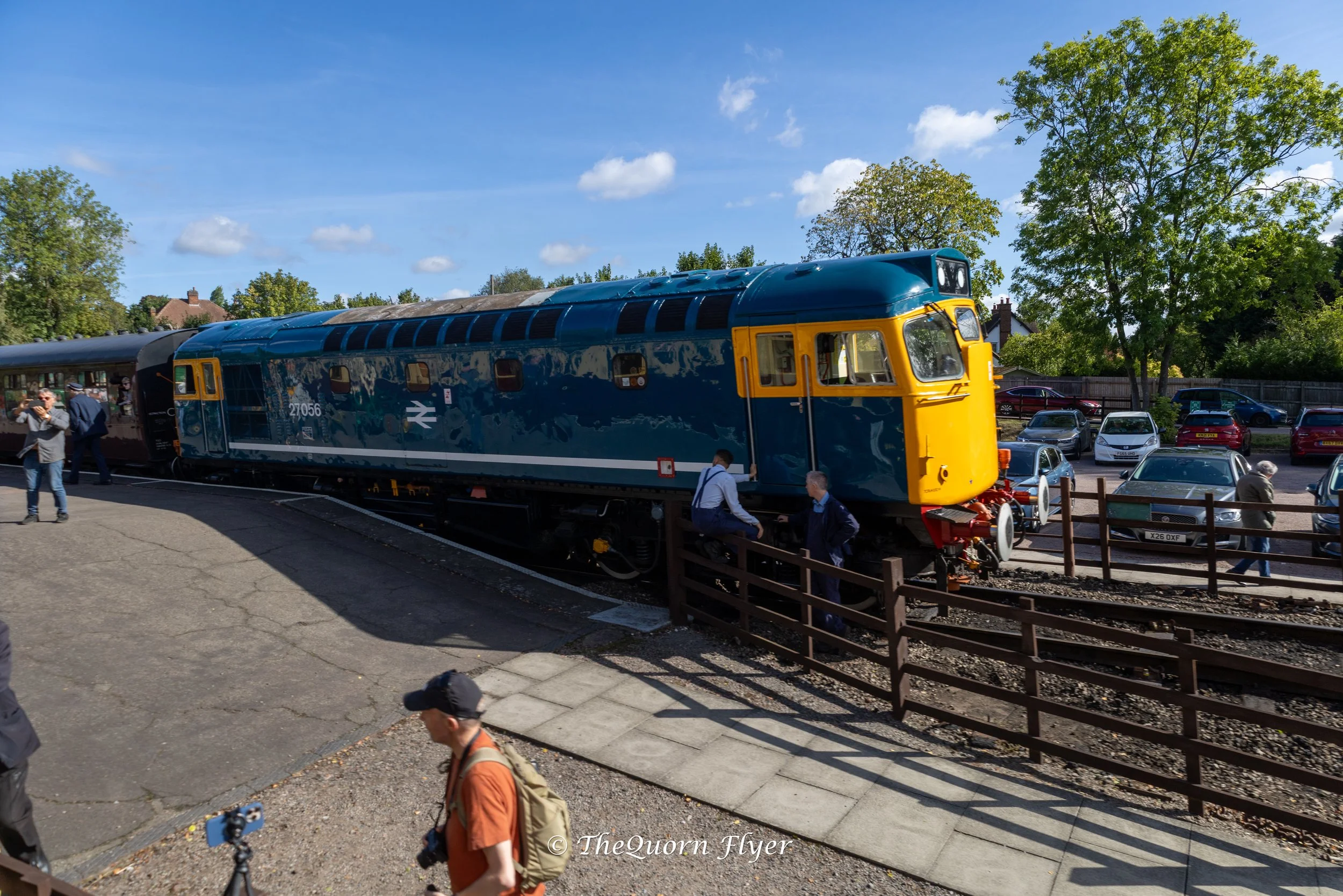 A vintage blue and yellow train parked outside on a bright, sunny day with trees and parked cars in the background. People are walking and sitting near the train.