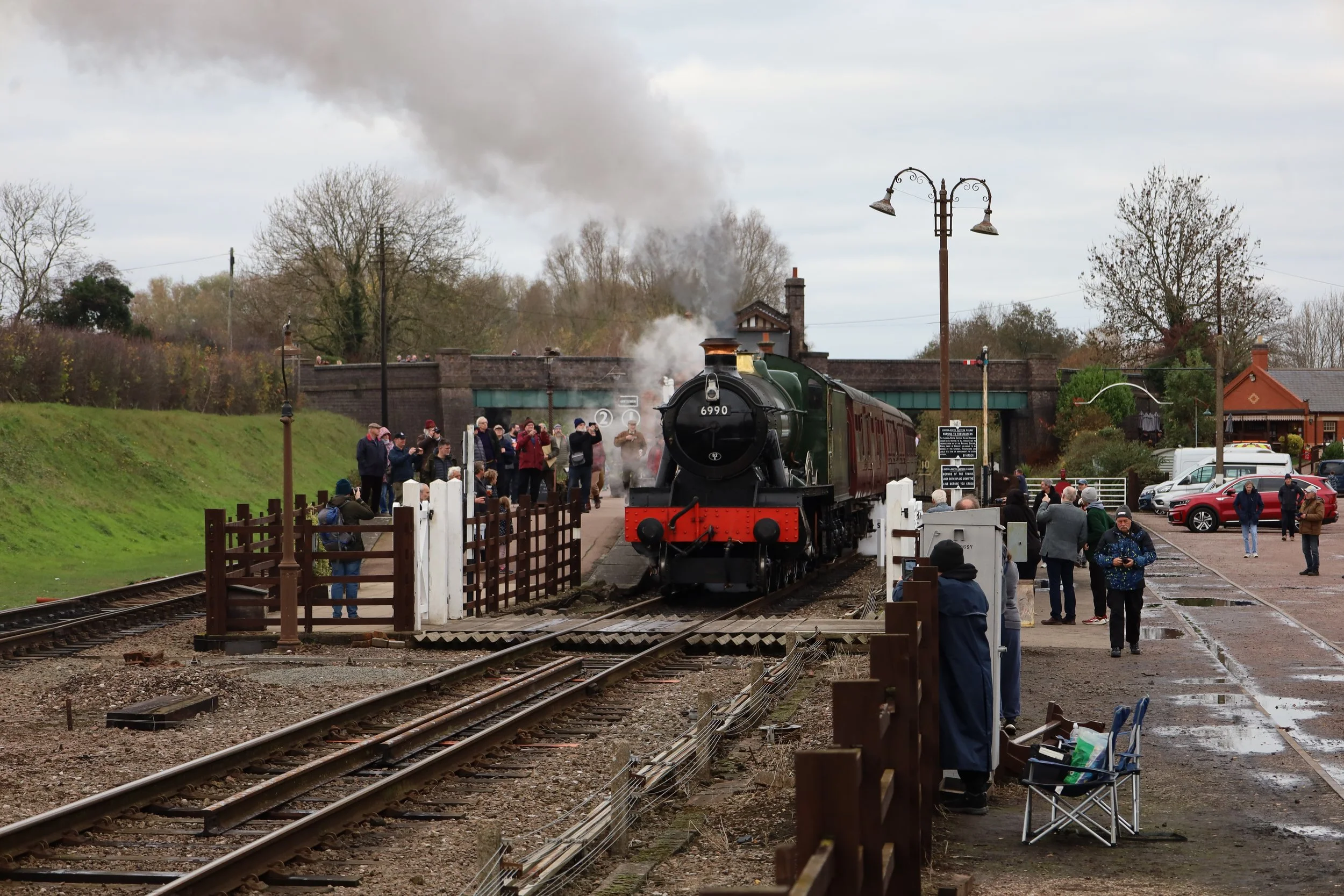 A vintage steam locomotive preparing to depart a train station on a cloudy day, with a crowd of people gathered along the platform, some taking photos, and vintage-style light poles and railway tracks in the background.
