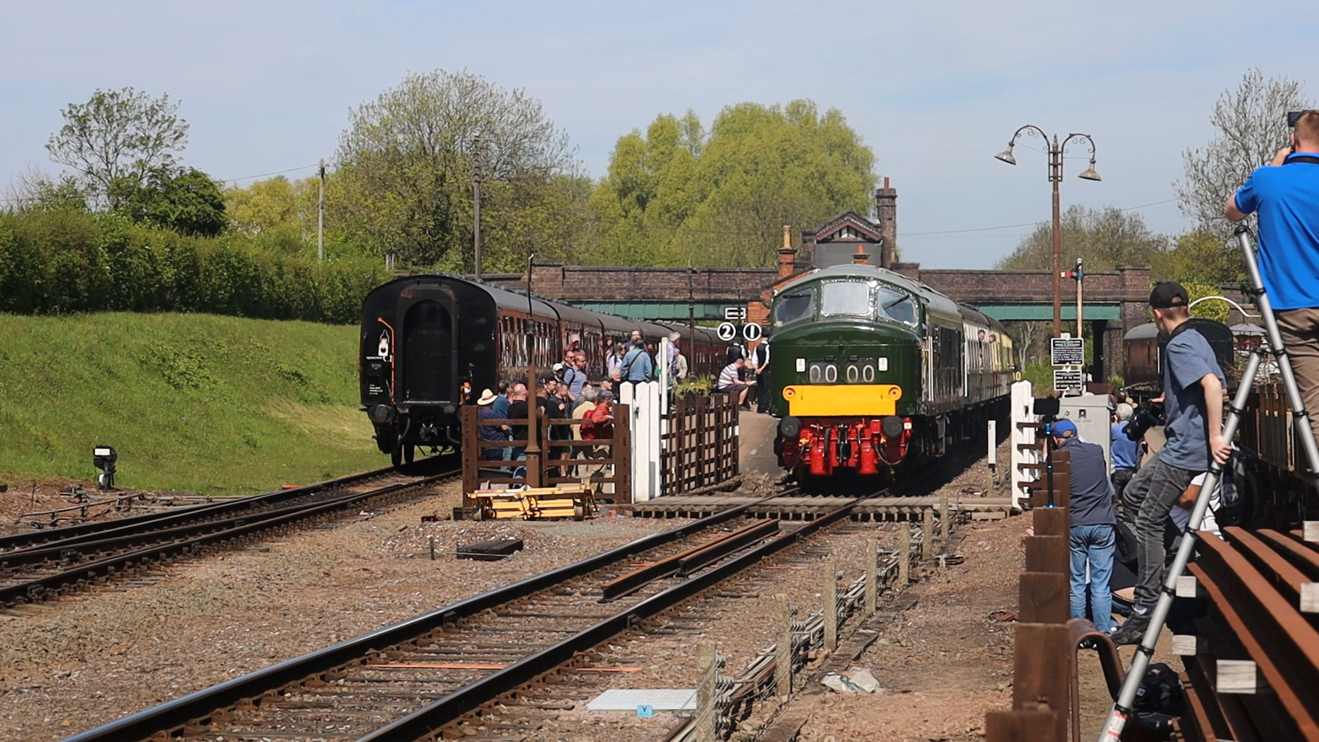 A vintage train station with a green and yellow train and black train cars, surrounded by people including photographers, in a rural setting with trees and a bridge in the background.