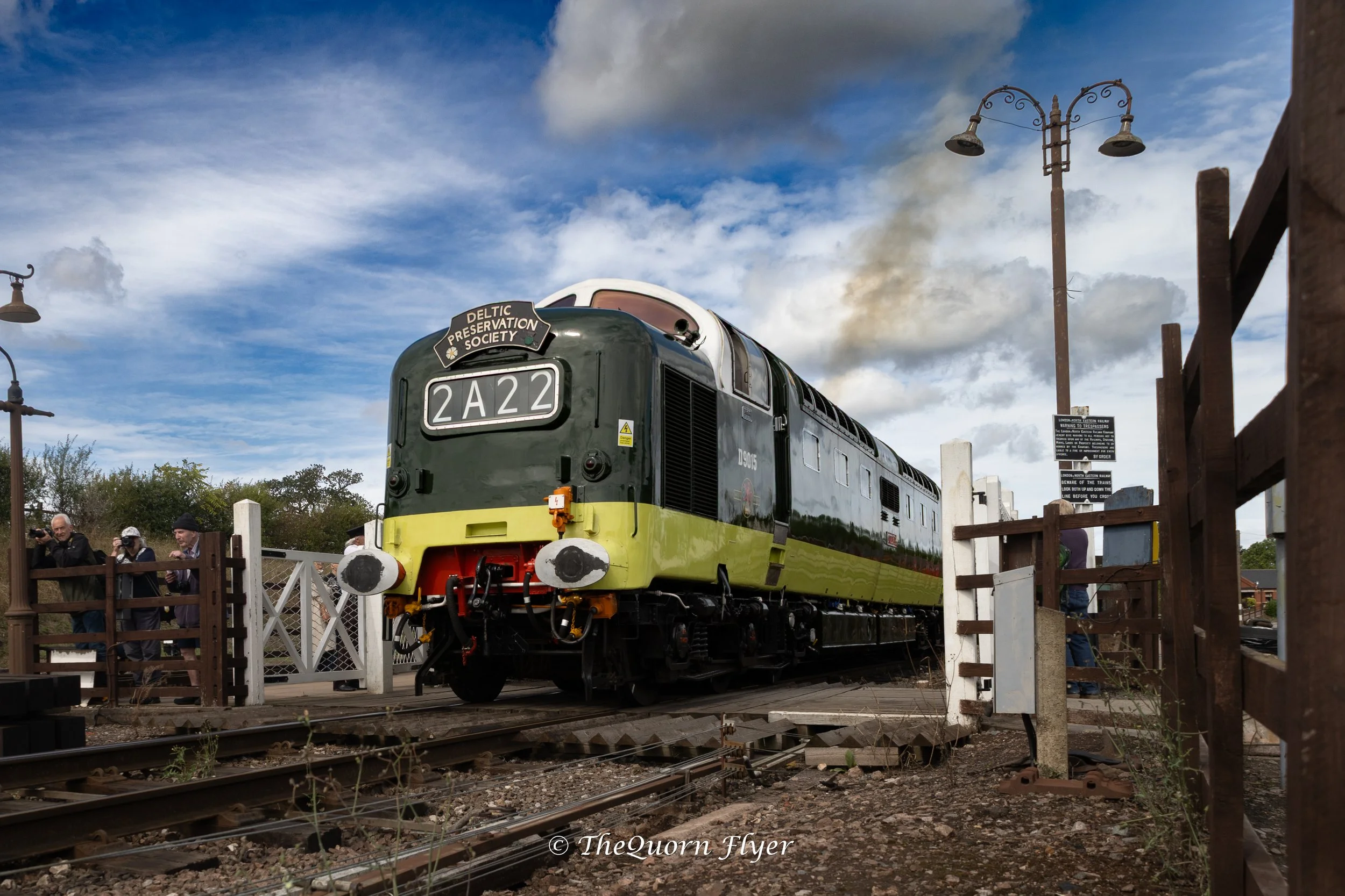 A vintage green and yellow train engine is on display outdoors, with a group of people watching nearby. The sky is partly cloudy with some smoke or steam rising in the background, and there are electric poles and warning signs around the train.