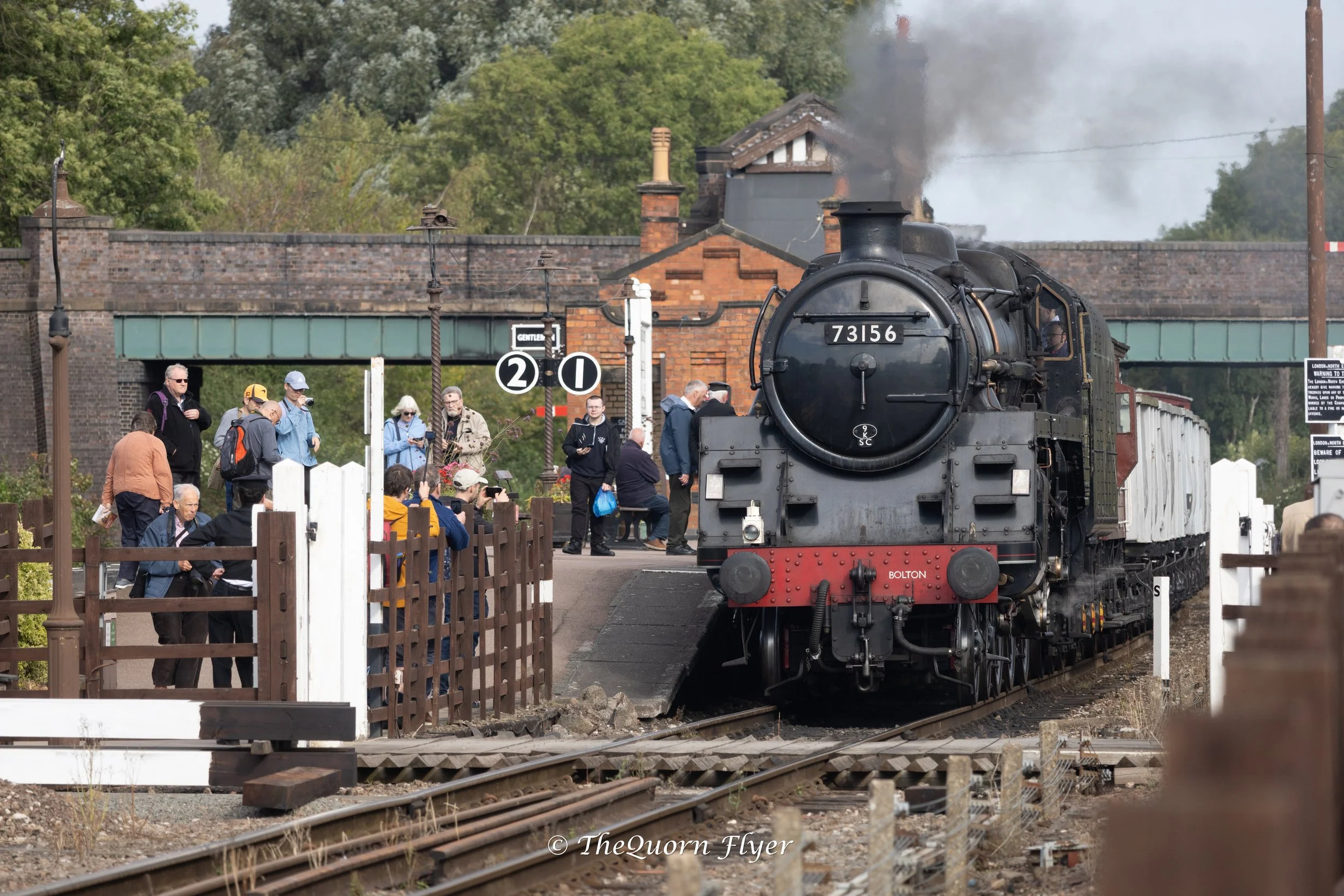 A vintage black steam locomotive with the number 73156 on the front, emitting steam as people gather around at a train station with trees and brick buildings in the background.