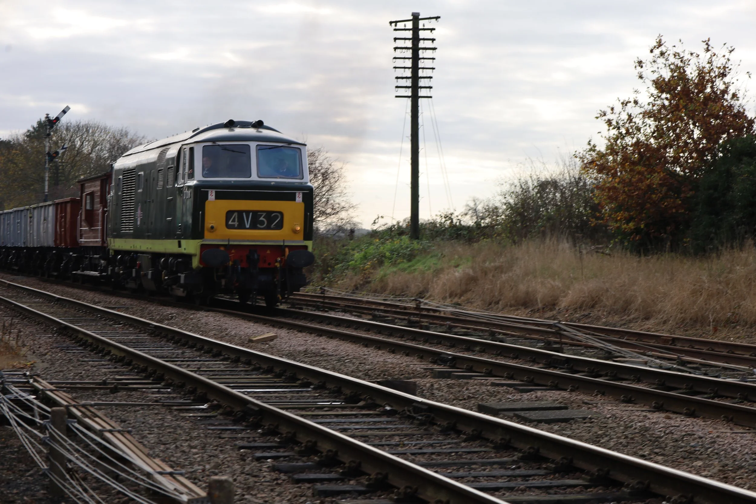 A vintage diesel train traveling on railway tracks through a rural area with trees and utility poles under a cloudy sky.