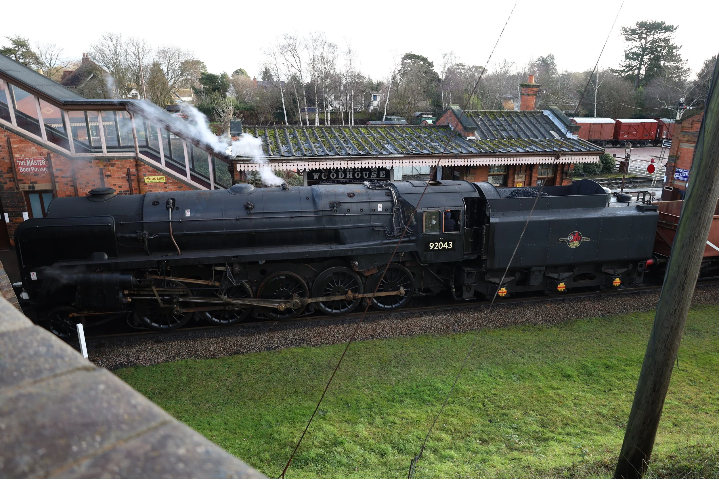 A vintage steam locomotive train parked at a station with a building labeled 'Woodhouse' and a sign that reads 'The Master Bootpolish'. The train has black paint and is emitting steam. Surrounding the station are trees and residential houses.