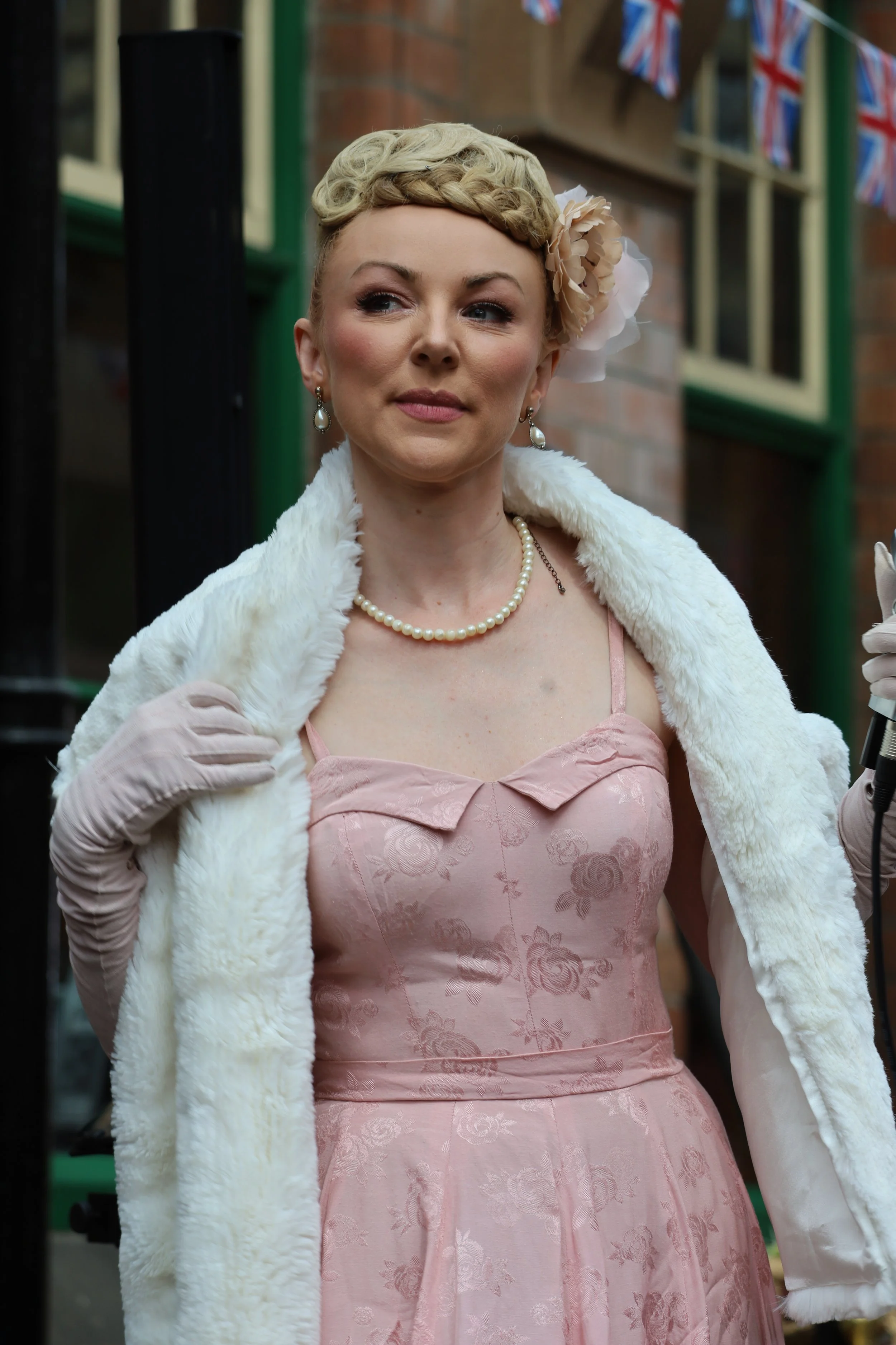 A woman with vintage-styled blonde hair wearing a blush pink dress with floral pattern, pearls, and pearl earrings, holding a white faux fur stole and wearing white gloves, standing outdoors in front of brick building with Union Jack bunting.