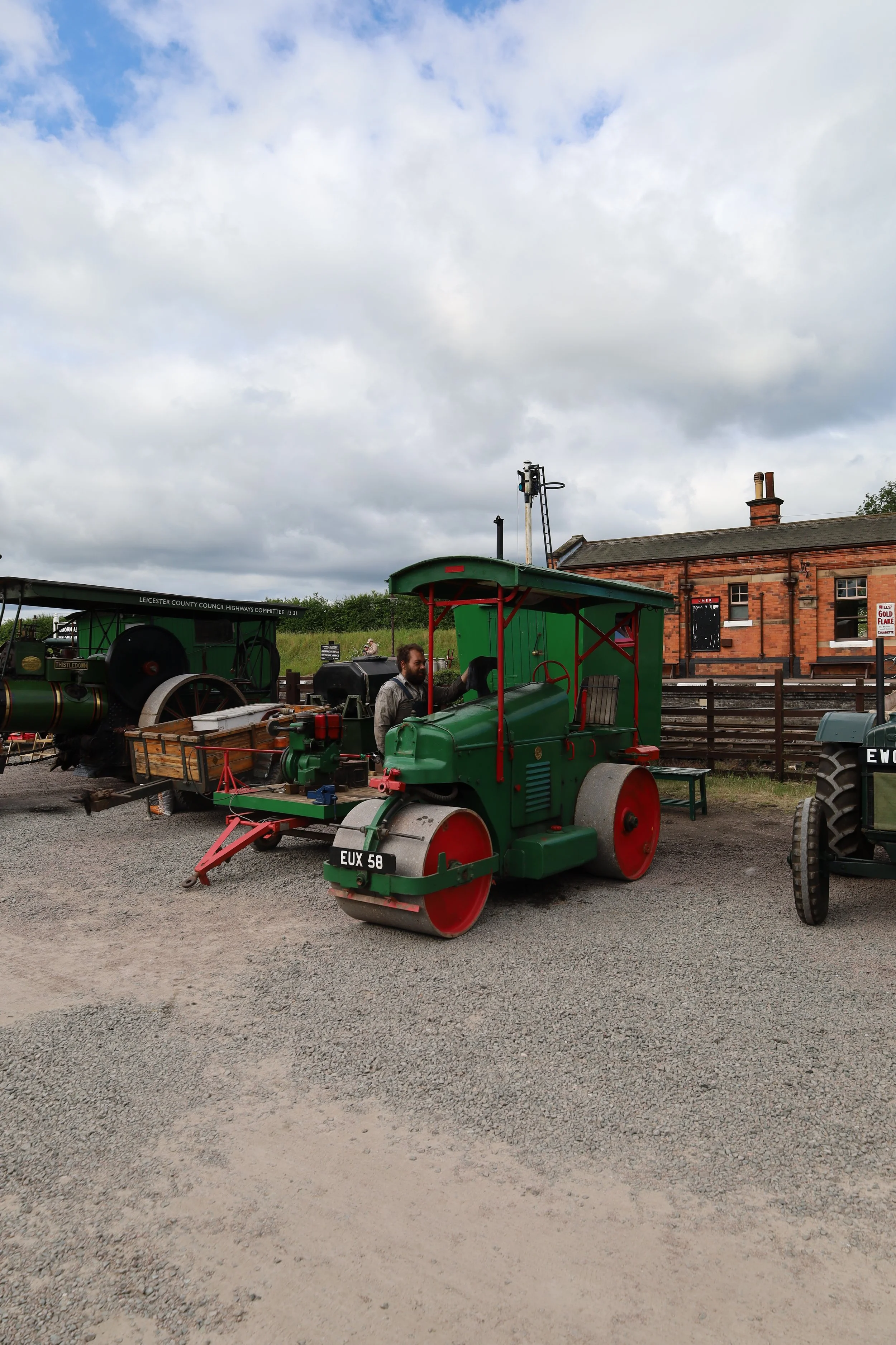 A vintage green steamroller at an outdoor historical train station or museum with a red brick building, a black vintage railroad cart, and a cloudy sky in the background.