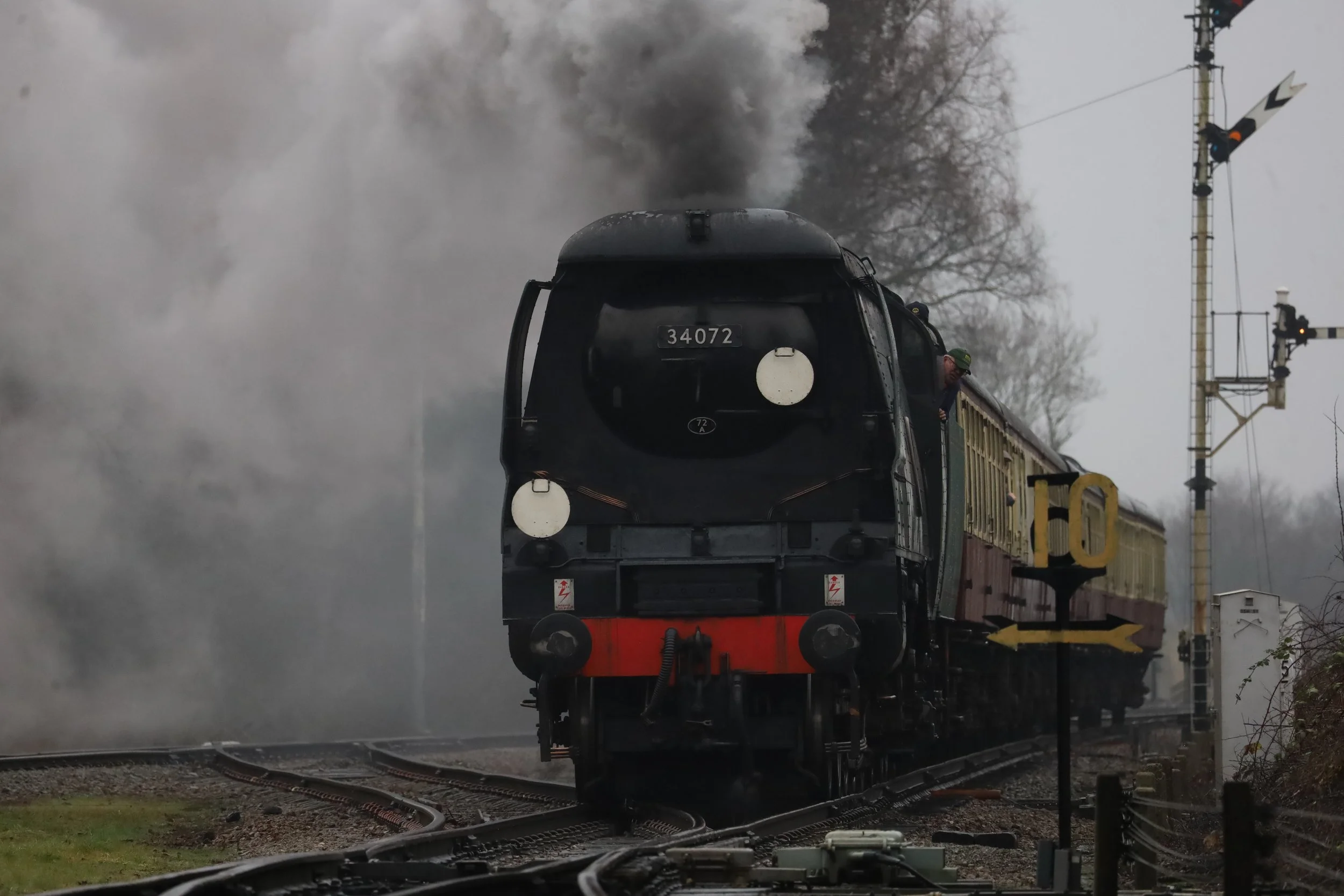 A black steam locomotive train emitting steam and smoke on railway tracks near a railroad crossing, with a person leaning out of the train window, and railroad signals visible in the background during daytime.