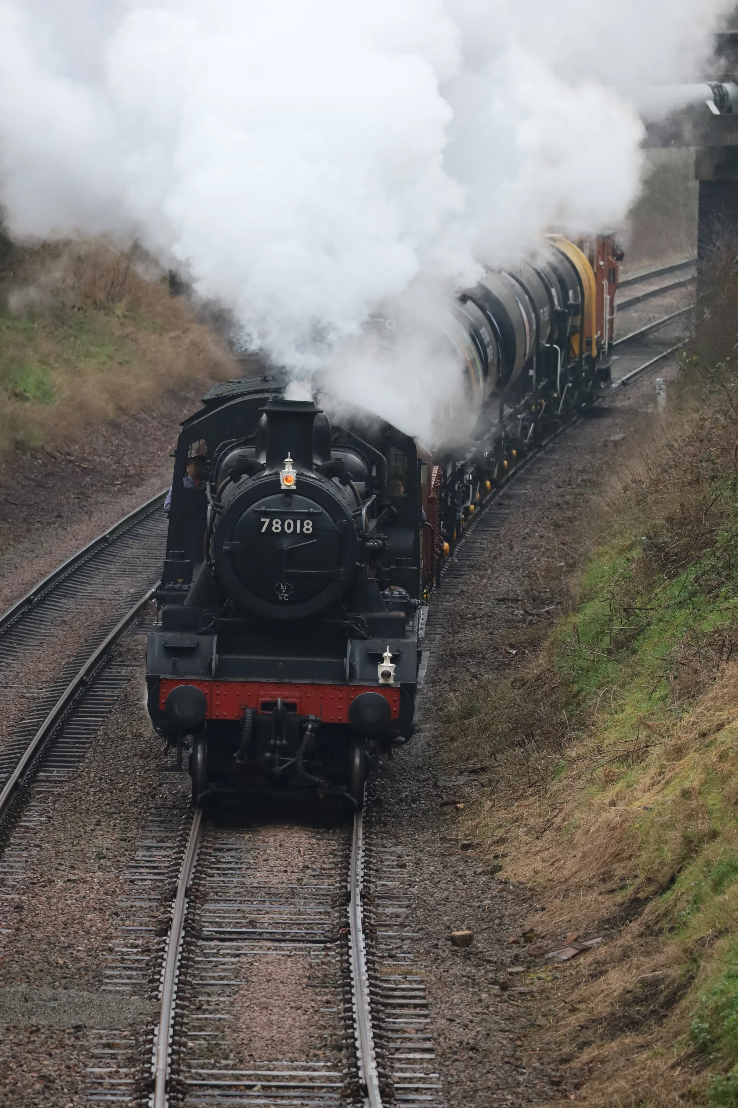 A black steam locomotive train with the number 78018 on the front, pulling several train cars through a rural area with grass and trees on the sides, emitting white steam from its chimney.
