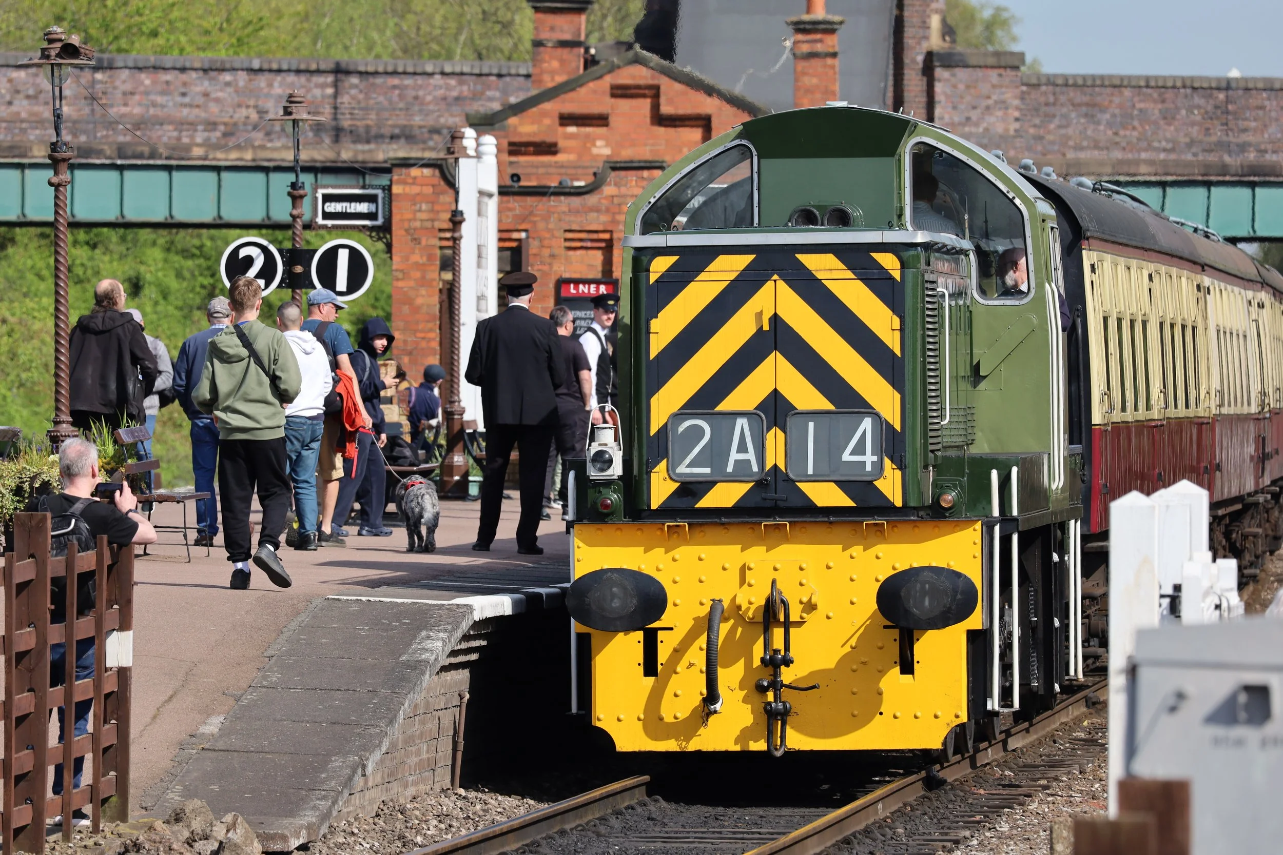 A vintage green train with yellow and black stripes on the front, labeled with the number 2A14, is at a train station platform. People are waiting around, some taking photos, with a few passengers visible inside the train. The background includes a brick building, a sign reading 'Gentlemen,' and greenery.