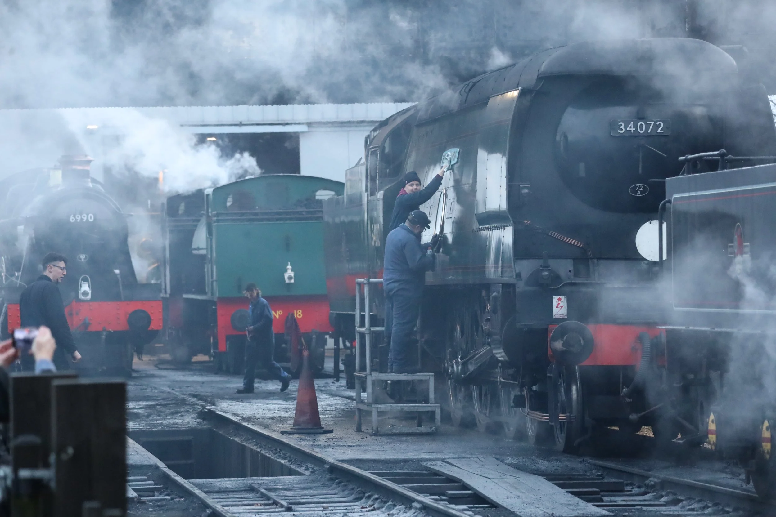 People cleaning and maintaining a large black steam locomotive train, with smoke around, under a bridge, and a person cleaning the side of the train