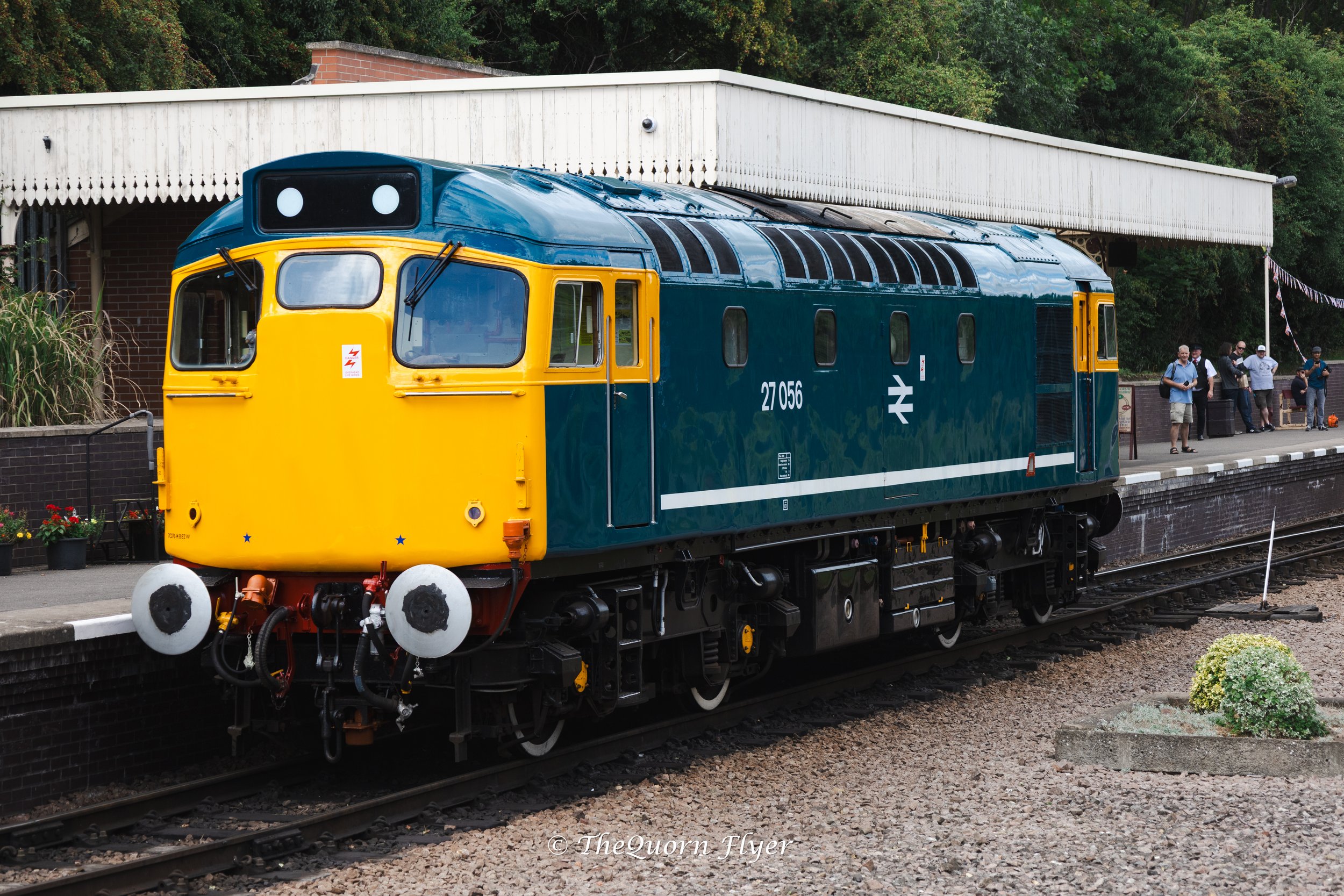 A vintage diesel train painted in dark green and yellow, stopped at a platform with a small group of people waiting nearby. There are trees in the background and some decorative flags.