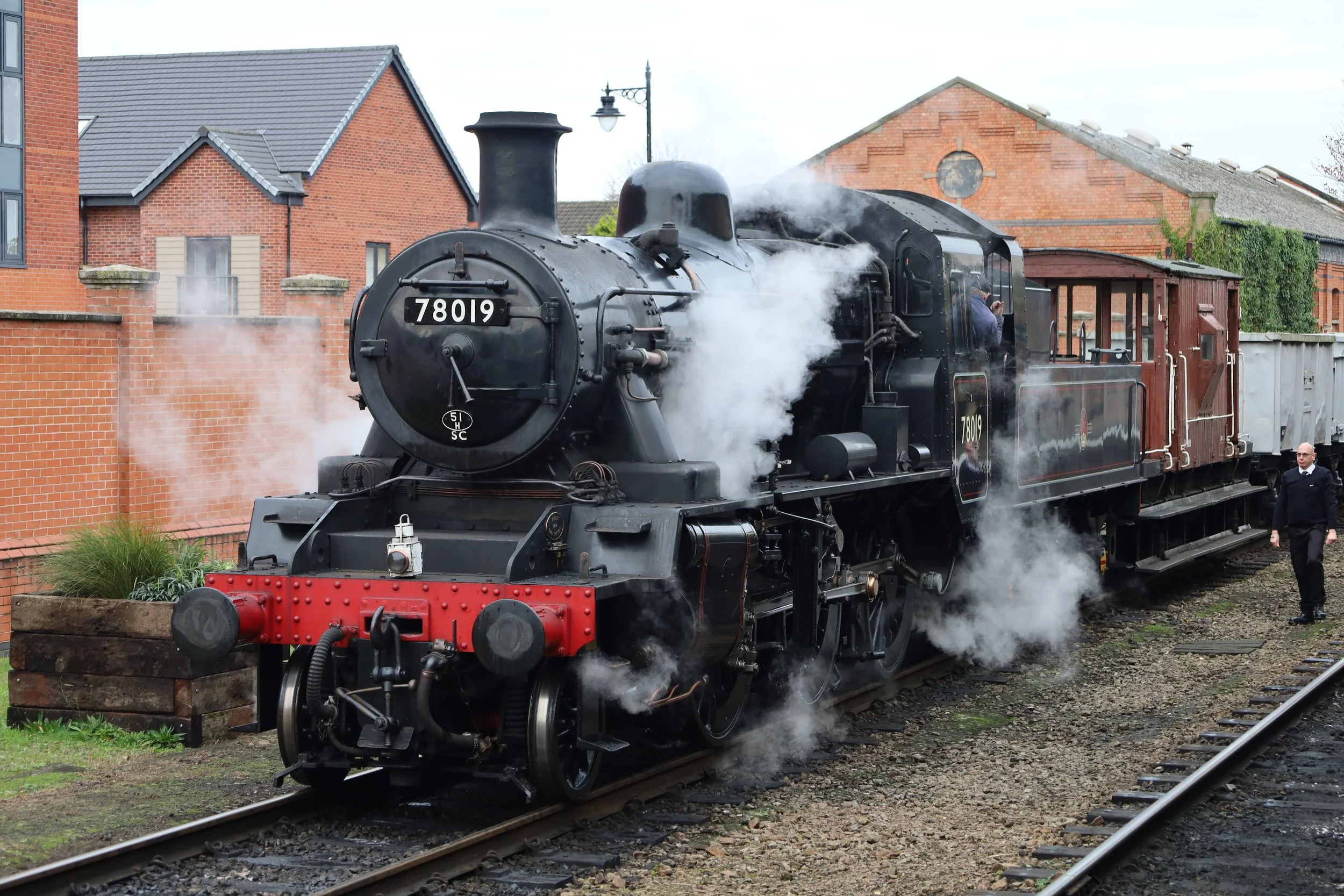 A vintage black steam locomotive with the number 78019 on the front, emitting steam, on a railway track in front of brick buildings and a street lamp, with a person in a suit walking nearby.