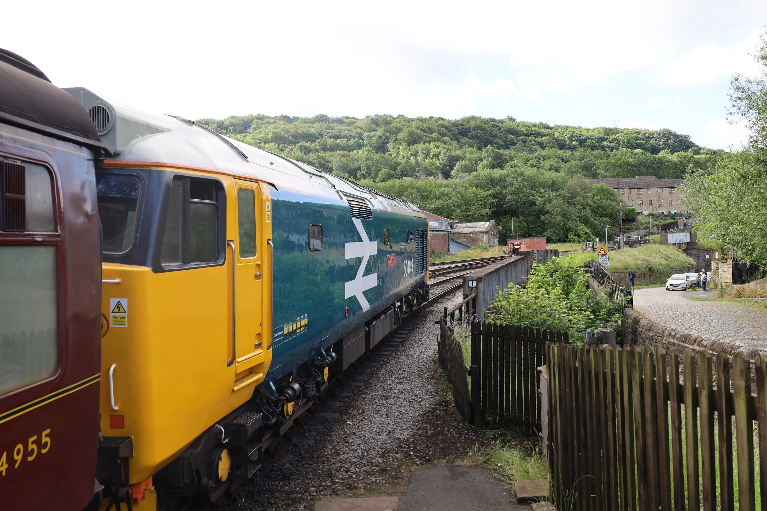 Part of a colorful train with a blue, yellow, and maroon color scheme on railway tracks in a rural area with trees, houses, and a hill in the background.