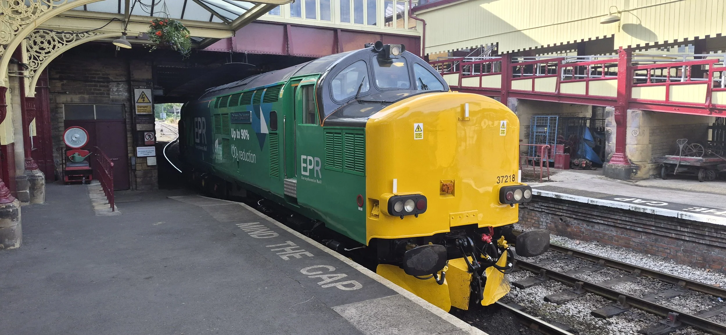 A colorful train with green and yellow colors and the letters 'EPR' and 'Europhoenix Rail' on its side, parked on tracks under a covered station platform with vintage-style architecture.