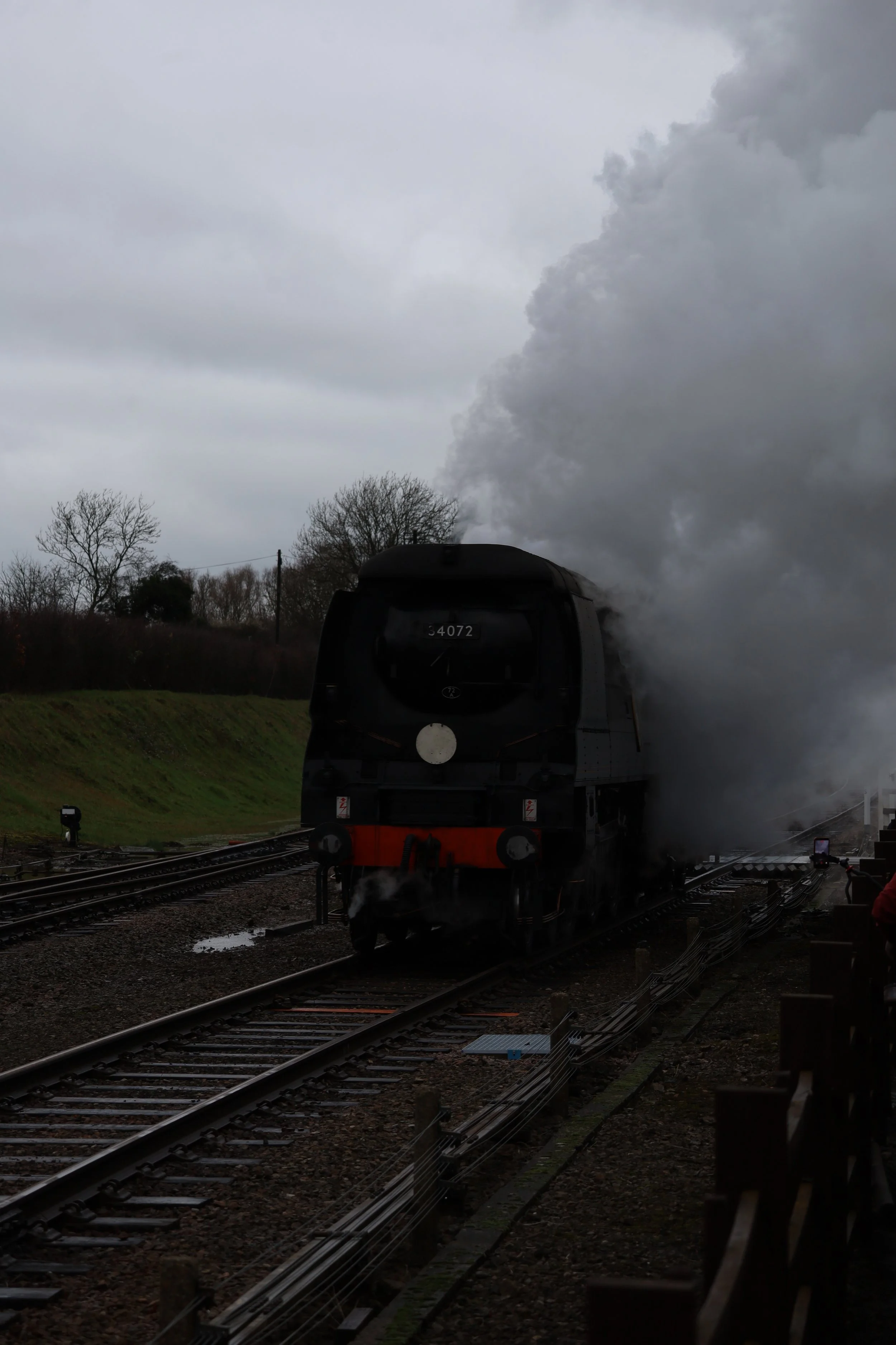 Black steam locomotive on railway tracks emitting a large amount of dark smoke under a cloudy sky, with leafless trees in the background.