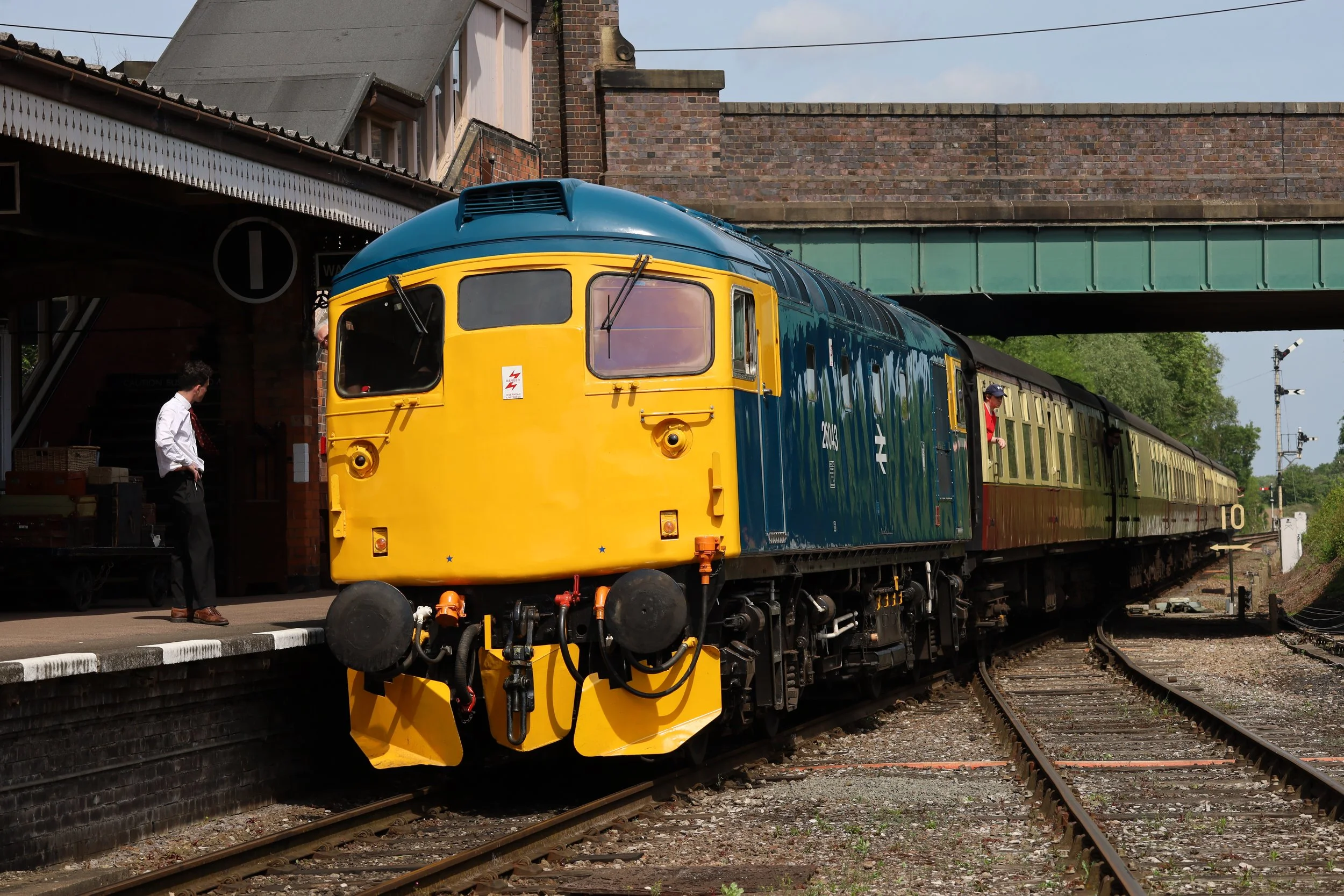 A vintage train with a yellow and blue front is stopped at a train station, with two men standing nearby and a bridge overhead.
