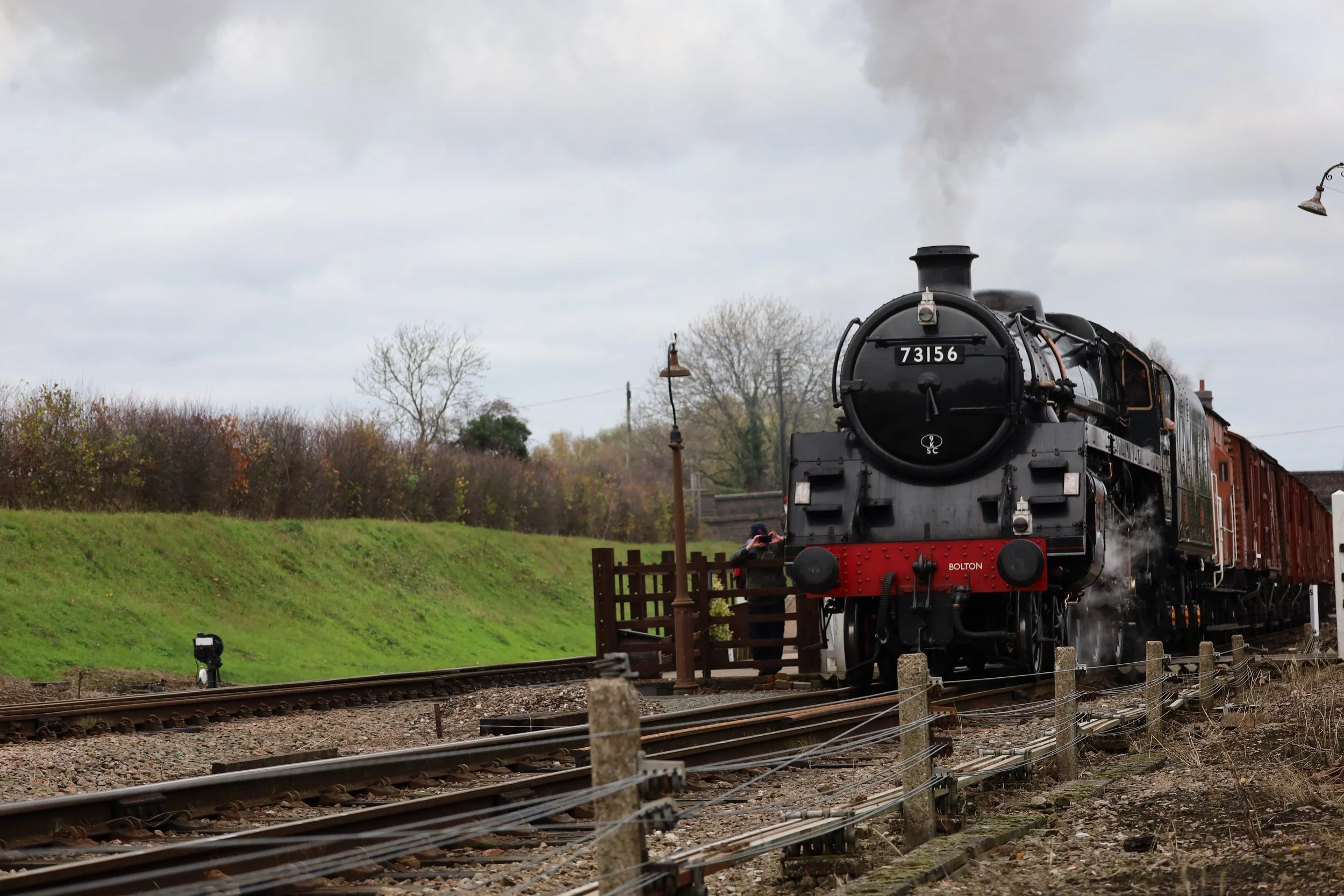 A vintage black steam locomotive with the number 73156 on the front, emitting smoke, crossing over tracks with a person taking photos on the side, and a cloudy sky overhead.