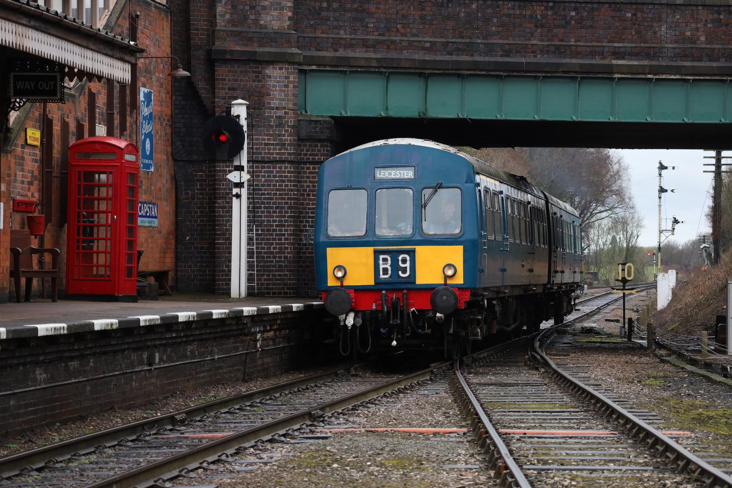 A blue vintage train identified as B9 exiting a railway station beneath a green bridge, with a red British telephone booth and brick station building on the platform, along with a signal light showing a red signal and tracks with switch points.