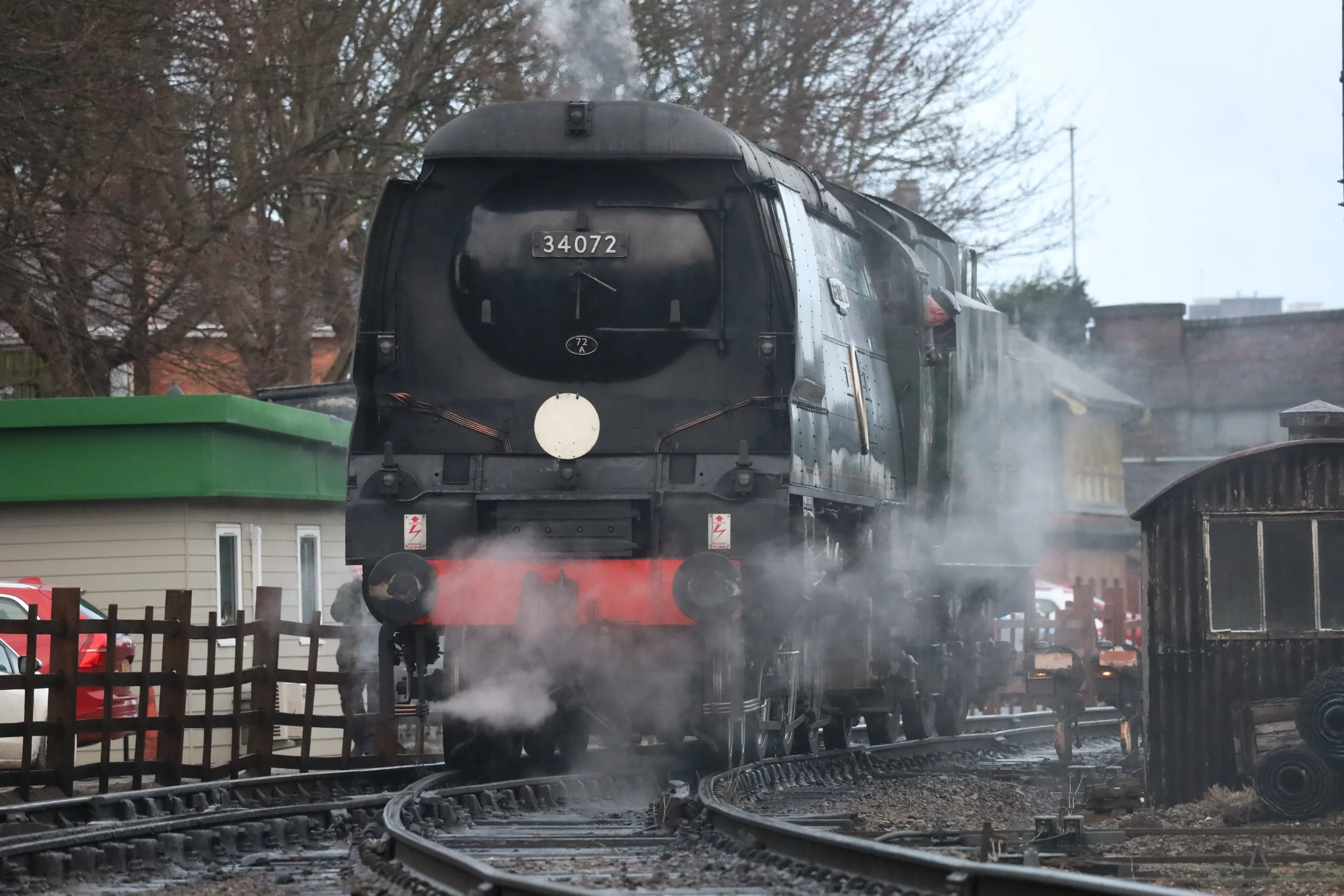 A black steam locomotive on railway tracks emitting steam, with a green building in the foreground and trees in the background.
