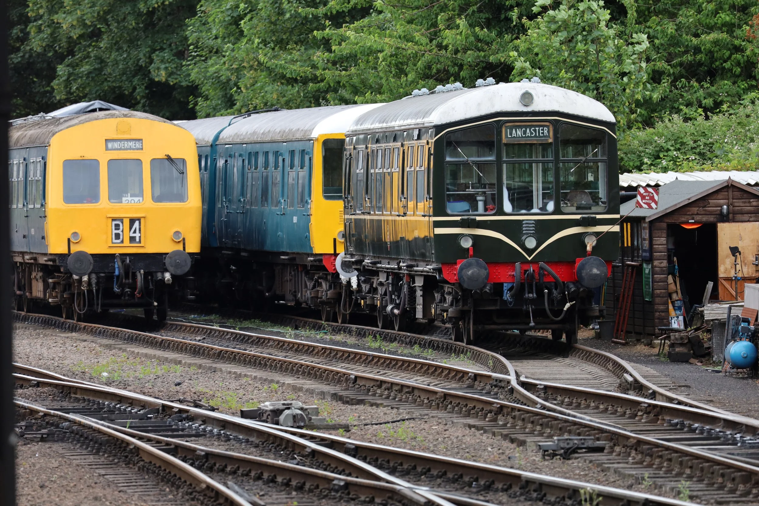 Vintage train cars on tracks in a wooded area, with a sign reading 'Lancaster' on one car and 'Windermere' on another.