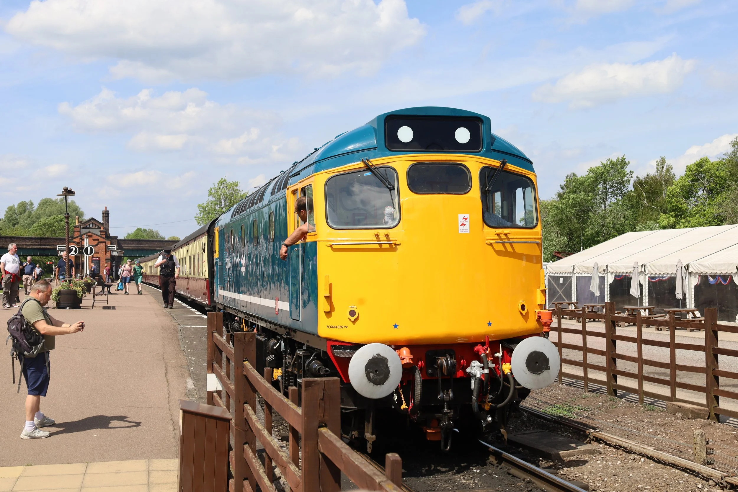 Colorful vintage train on display at a train station, with people walking around and a large white tent in the background under a partly cloudy sky.
