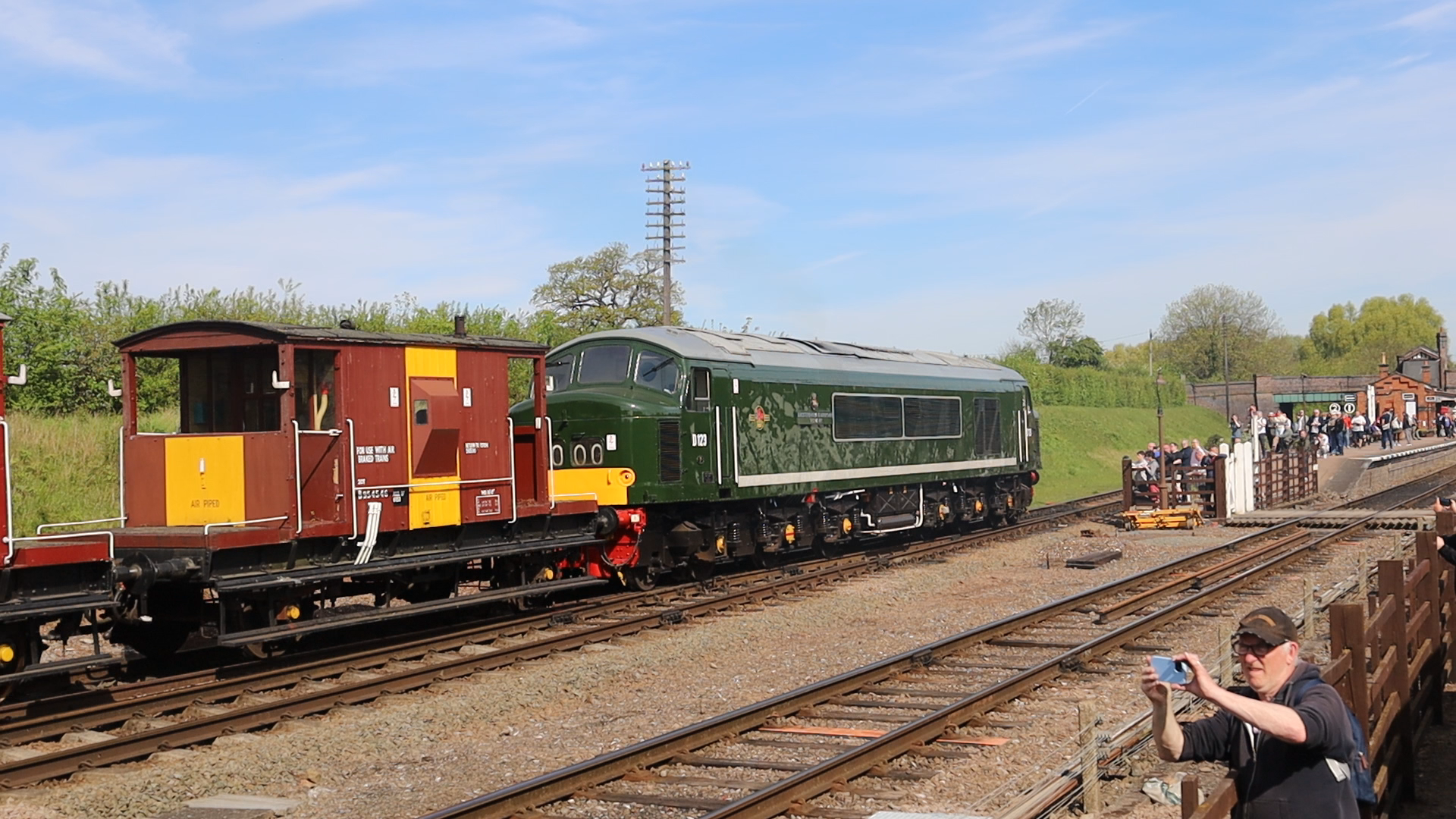 A green vintage train with a yellow front is on the tracks at a train station with people waiting on the platform and taking photos. The sky is blue with some clouds and trees are in the background.