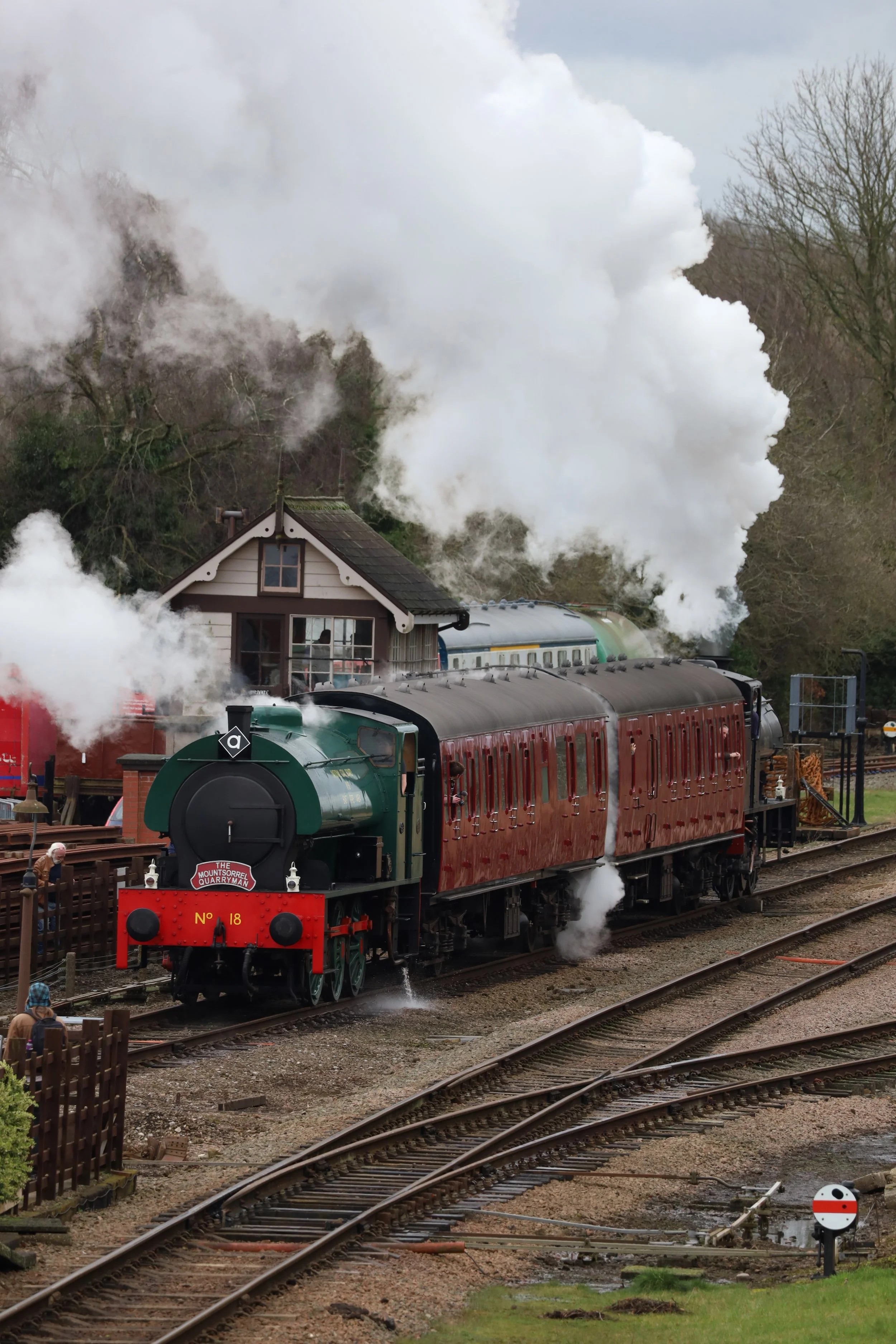 A vintage steam train with red carriages and a green engine is emitting large clouds of white steam at a railway station, with people nearby and a small building in the background.