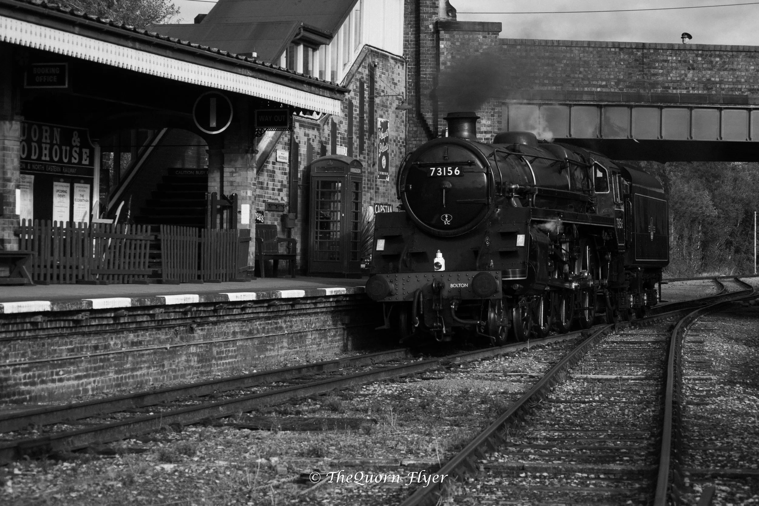 A black and white photo of a vintage steam locomotive train at a station, with brick buildings and platform area nearby, including a telephone booth and signage.