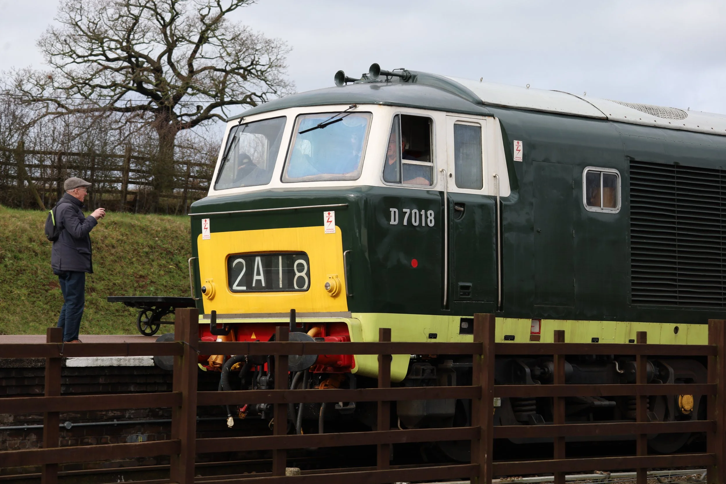 A man with a gray hat and glasses is standing outdoors near a vintage green locomotive with a yellow front, and he is taking a photo of it. The locomotive has the number D7018 on its side, and there are leafless trees and a wooden fence in the background. The sky is cloudy.