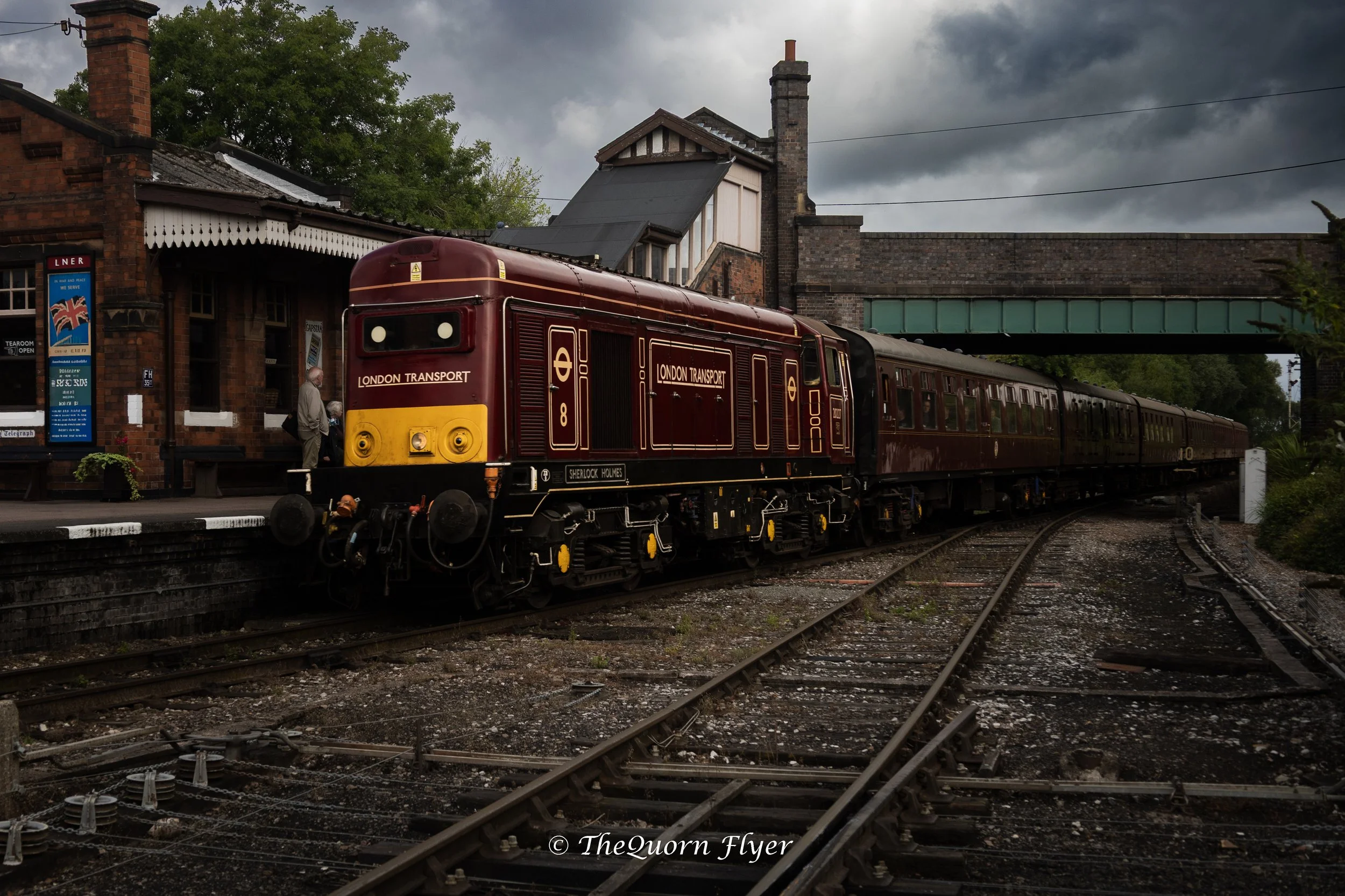 A vintage red and yellow London Transport train stopped at a station platform with a brick building and dark cloudy sky in the background.