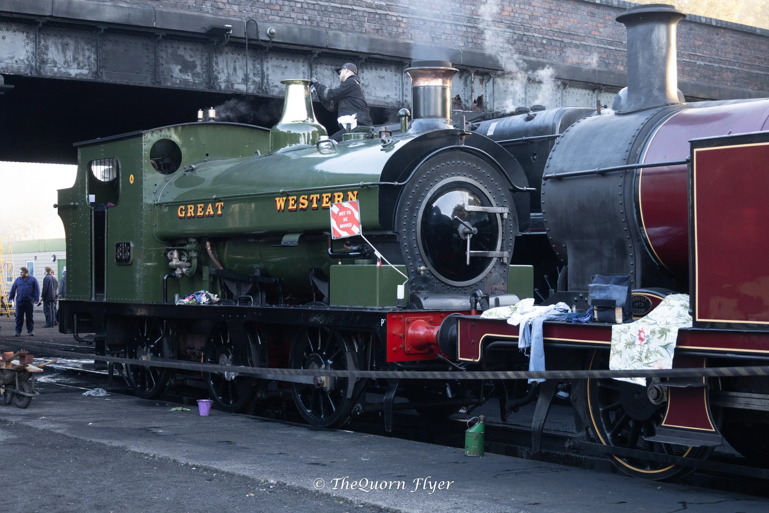 A vintage green and black steam locomotive named 'Great Western' is displayed outdoors, with a man working on it. The locomotive is near a brick wall, and there are people and objects like clothes and cans around it.