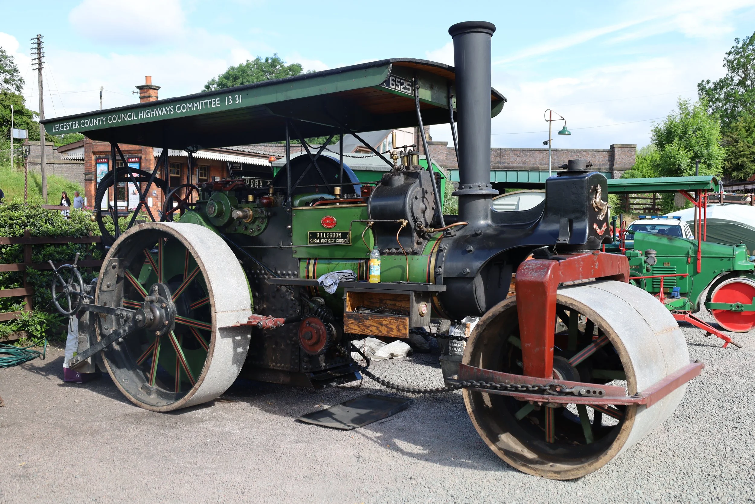 Vintage steam-powered tractor with large metal wheels, displayed outdoors at a fair or exhibit with greenery and a brick building in the background.
