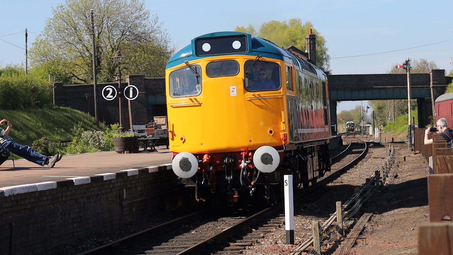A yellow train with a blue face is arriving at a train station platform. There are two people taking photos of the train, one sitting on the platform and one standing. Trees and a brick bridge are visible in the background, with train tracks running underneath.