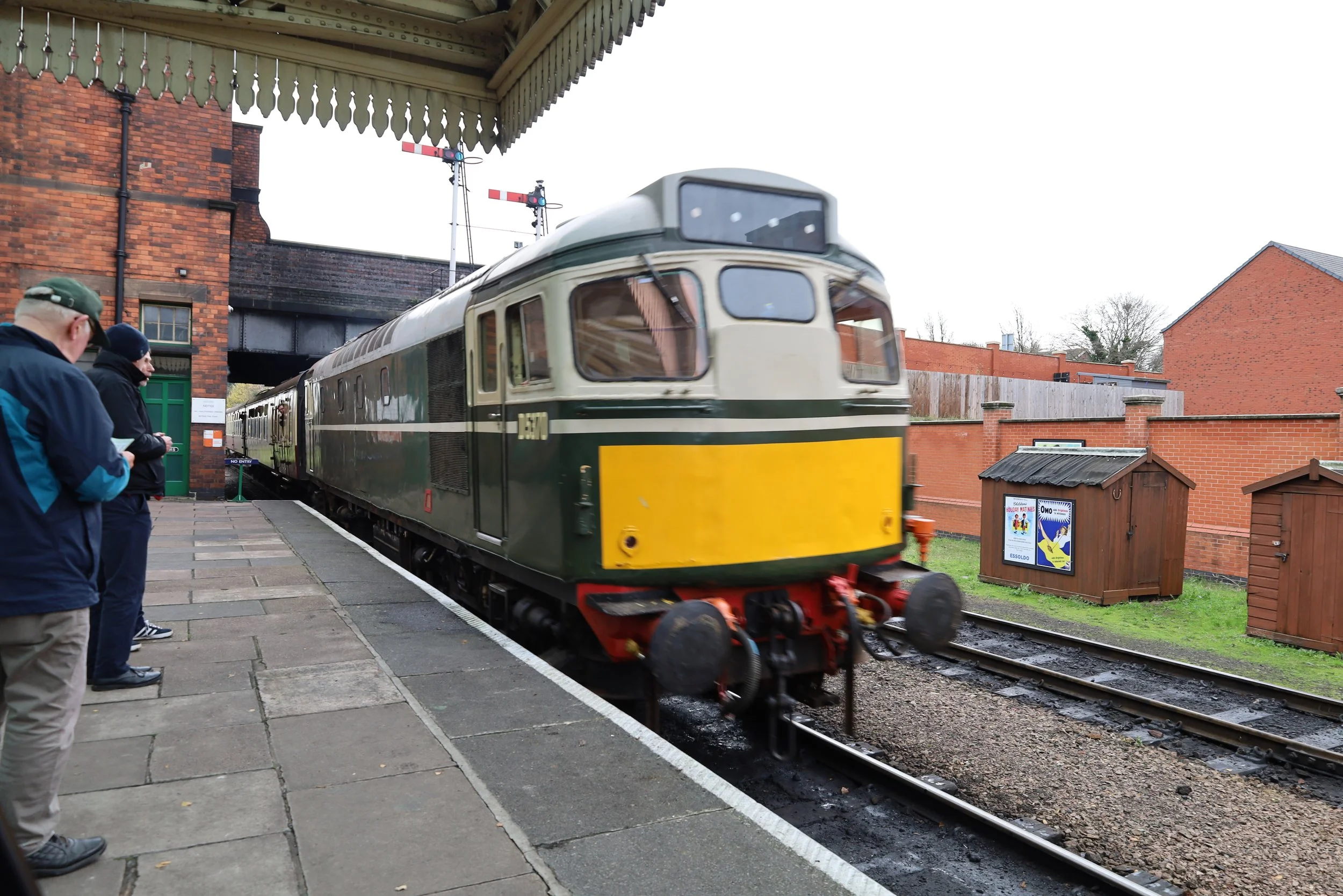 A vintage green and yellow train arriving at a station platform with three people waiting and looking at it. The platform and surroundings include brick buildings and a small wooden structure, with railway signals and bridge in the background.