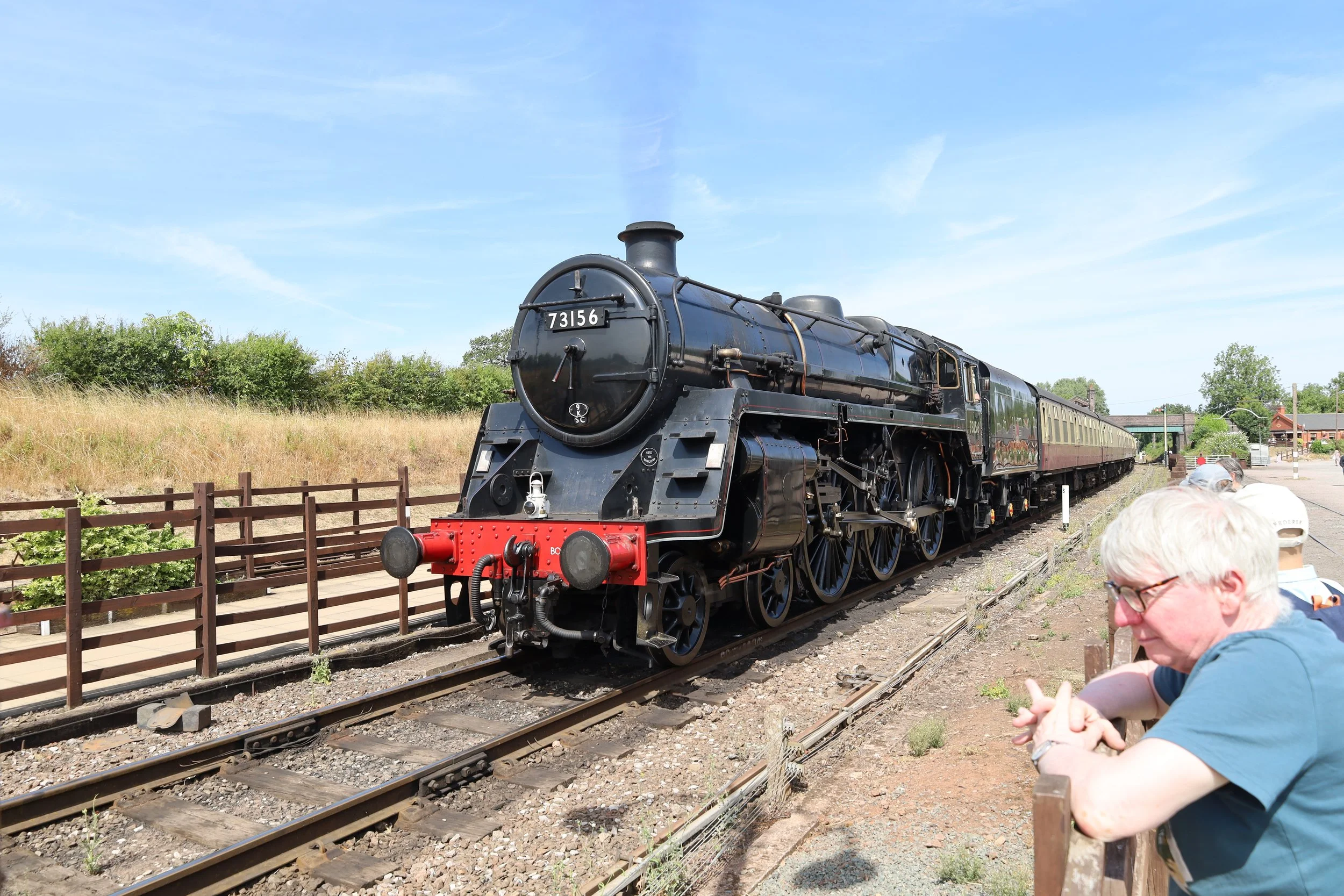 A vintage black steam train with the number 73156 on the front is on railway tracks outdoors. There are people, including an older woman in the foreground, looking on and taking photos, with a wooden fence along the tracks, grassy fields, trees, and a small building in the background under a blue sky.