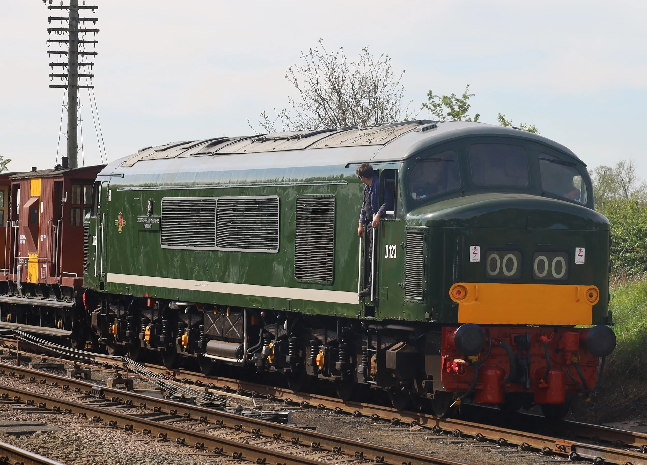 A green vintage train with a person leaning out of the window, on parallel railway tracks with electric poles in the background.