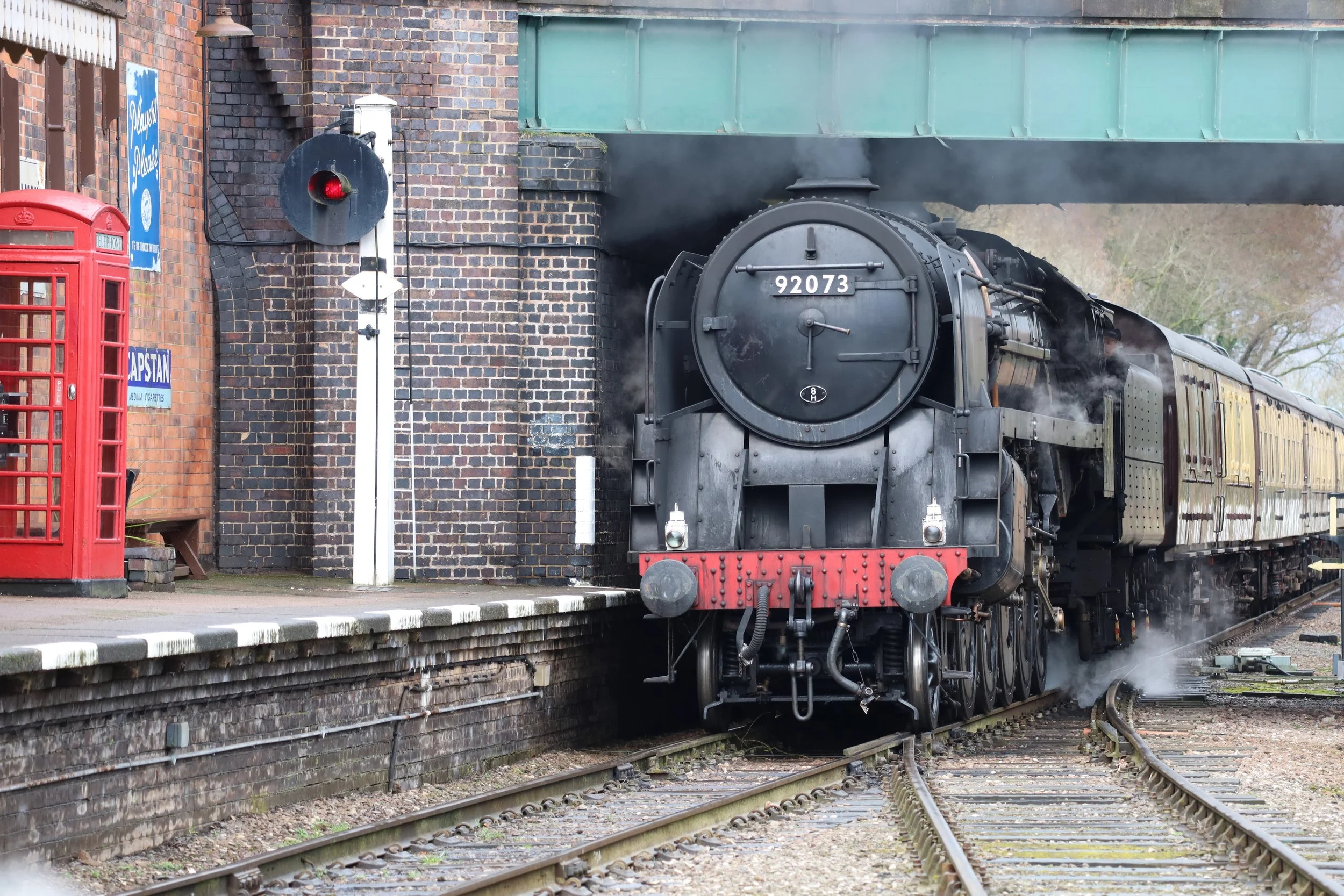 A vintage black steam locomotive with the number 92073 emerging from a brick train station under a green bridge, with smoke rising from its smoke stack. On the left side, there is a red British telephone booth and a blue sign on the wall.