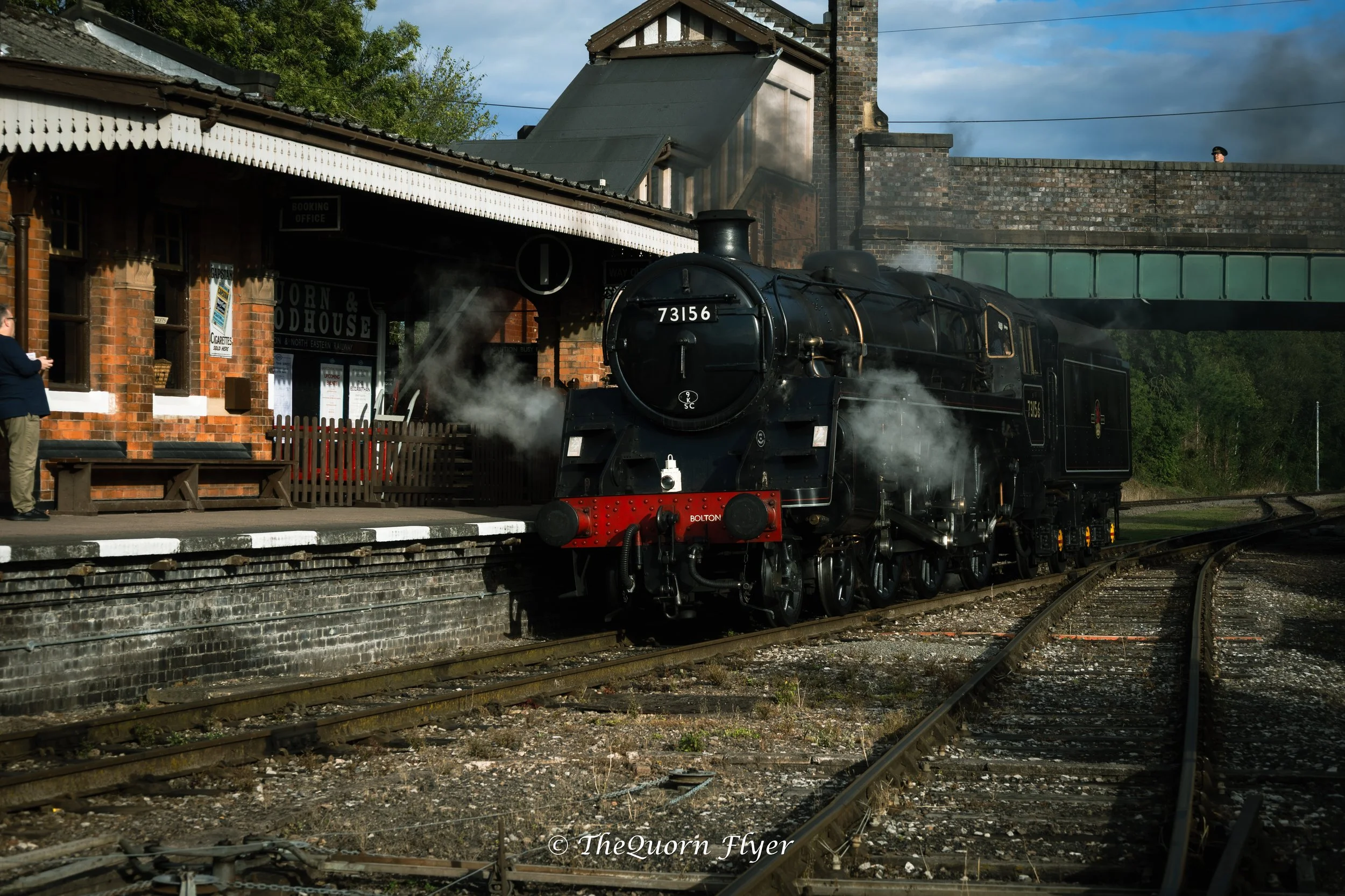 A vintage steam train with the number 73156 on its front, pulling into a station beneath a brick building with a covered platform. The station building has a booking office sign and a window poster. A person stands on the platform, and a person in a black hat is visible above the station wall. The train emits steam, and the sky above is partly cloudy.