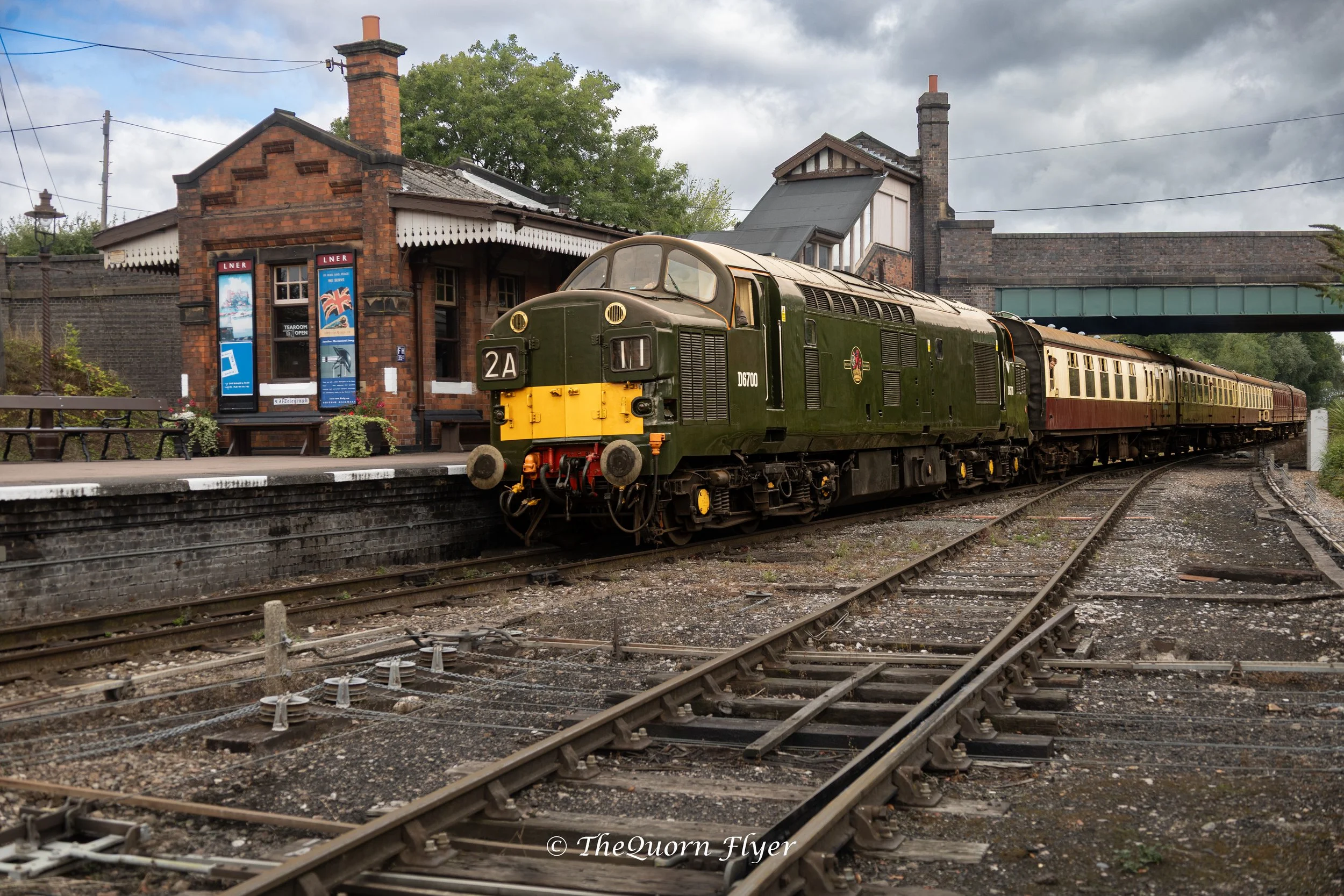 Vintage green train at station with brick building in background and cloudy sky.
