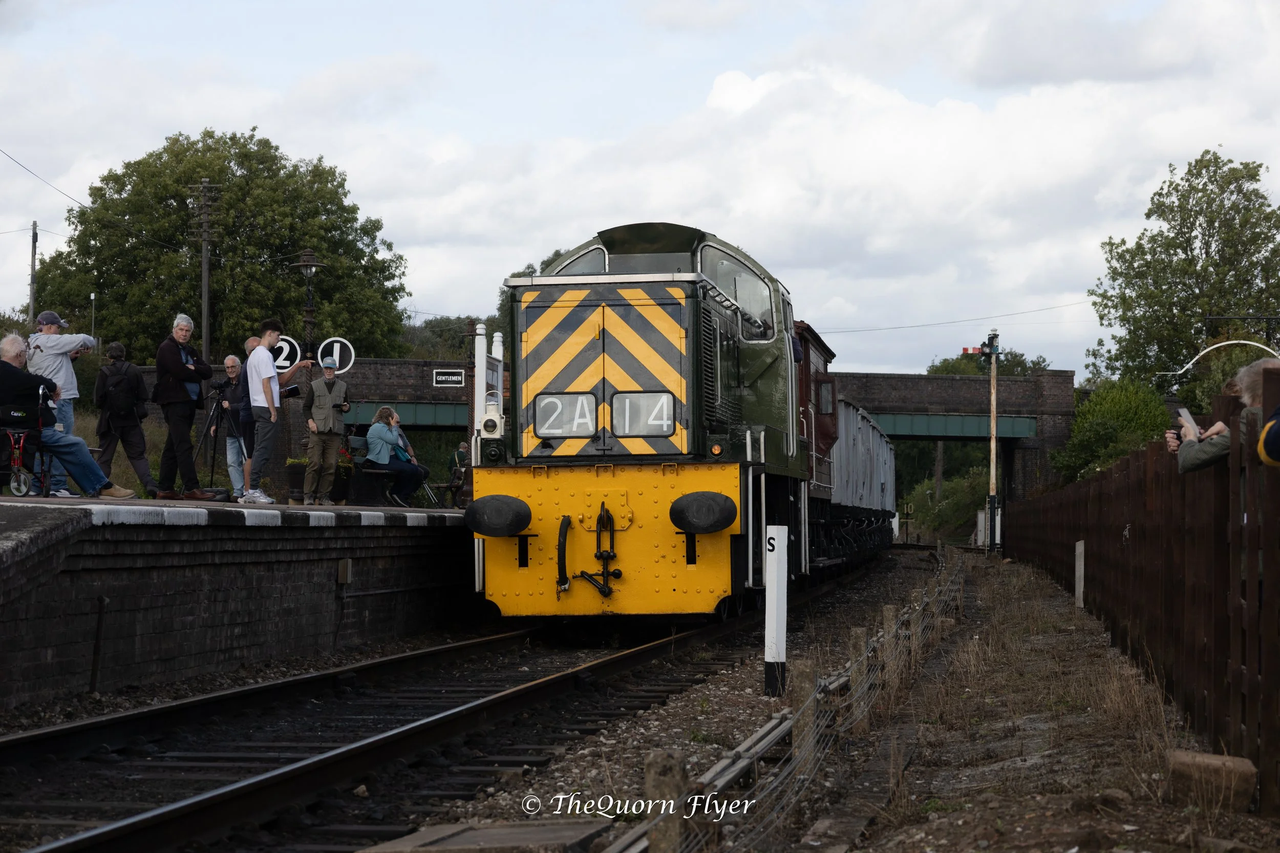 A vintage train with black and yellow warning stripes on the front, approaching a platform with people waiting, some taking photos. The scene includes trees, a bridge, and a cloudy sky.
