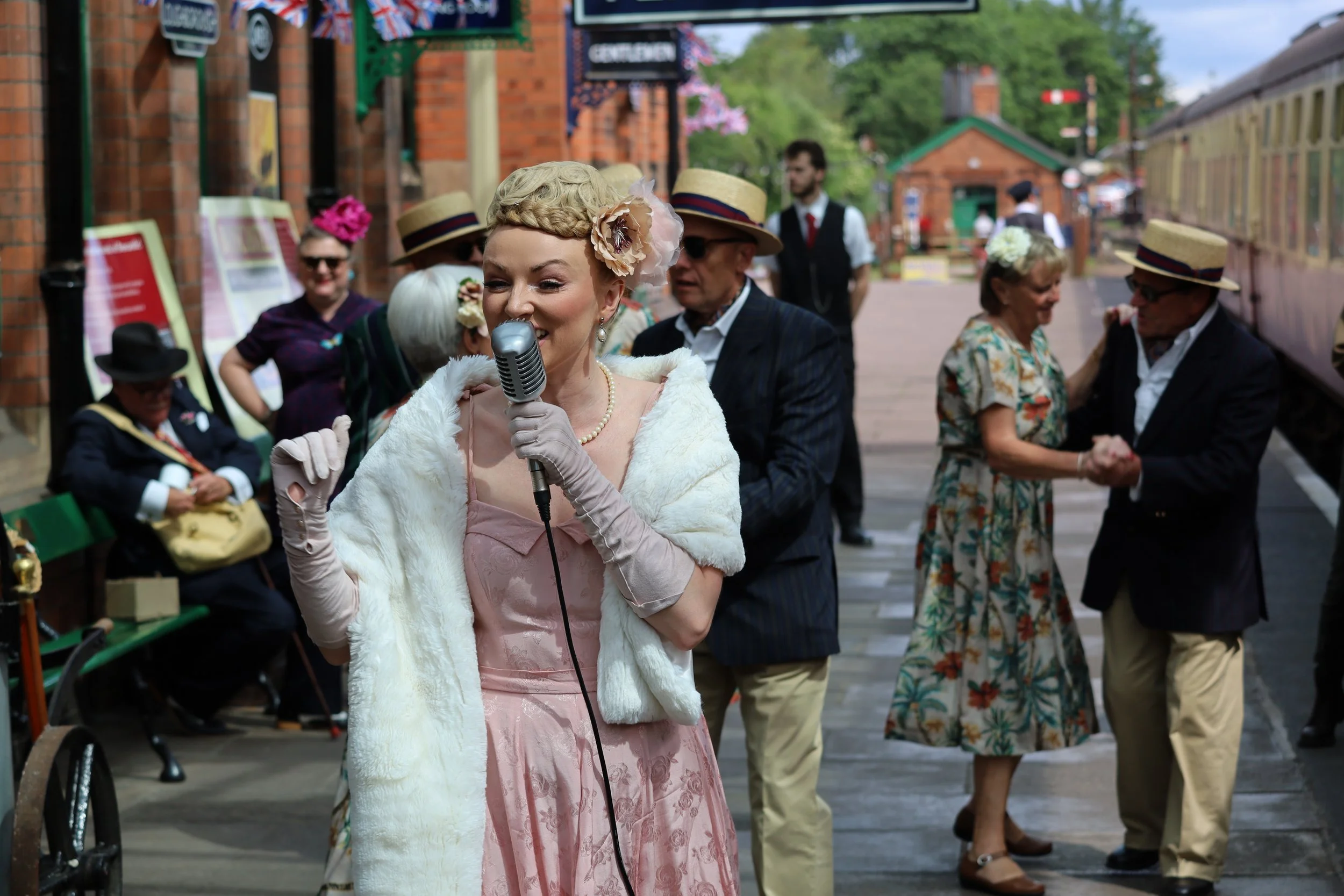 A woman dressed in vintage clothing with a pink dress and white fur stole, singing into a microphone during a street event, while people in period costumes dance and socialize in the background.