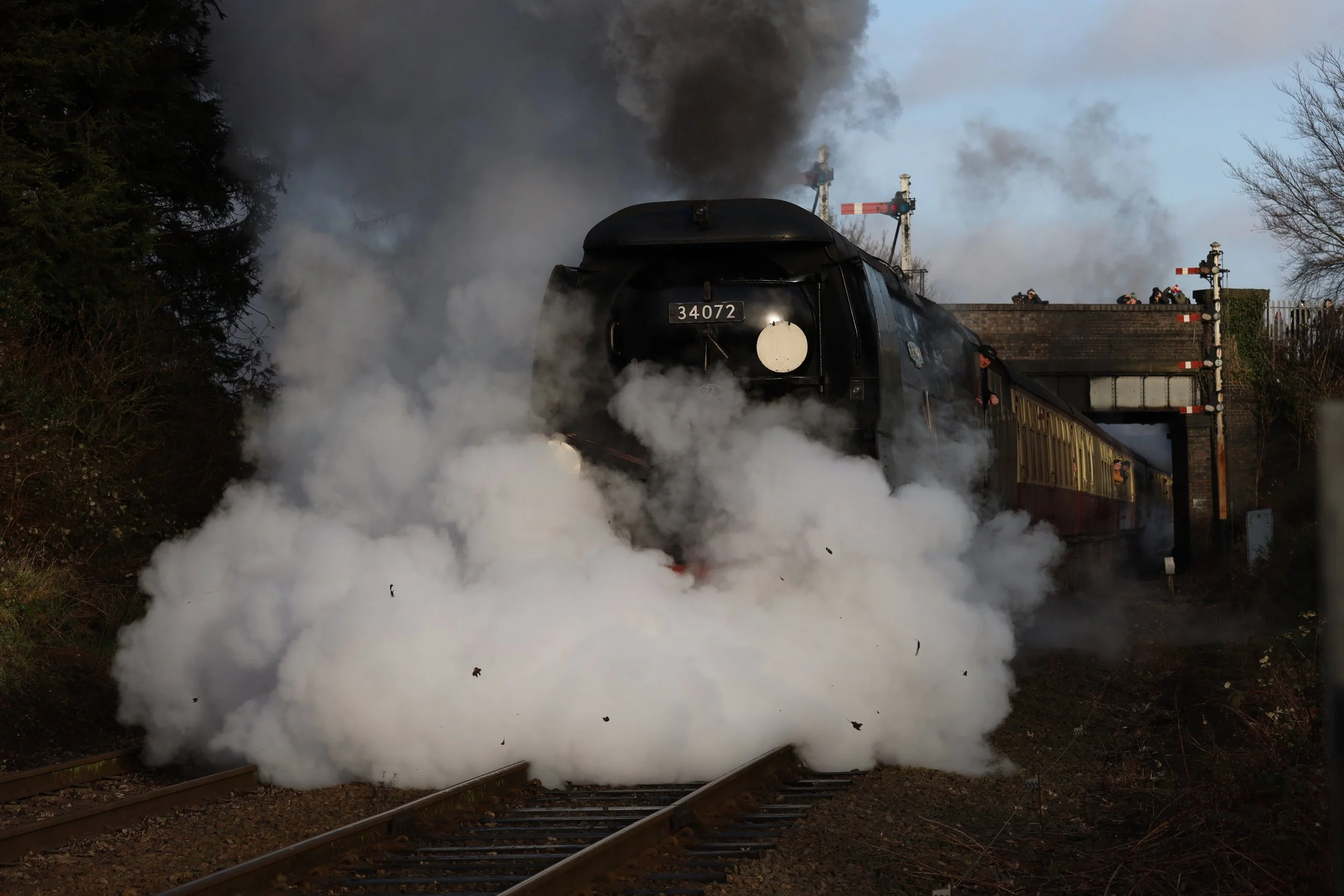 A vintage black steam locomotive with the number 34072 on the front, emitting thick black smoke as it crosses a railway bridge. The bridge is supported by brick pillars, and several people are visible on top of the bridge, possibly railway personnel or observers. The scene is framed by trees on either side of the tracks, with the ground covered in dirt and gravel.