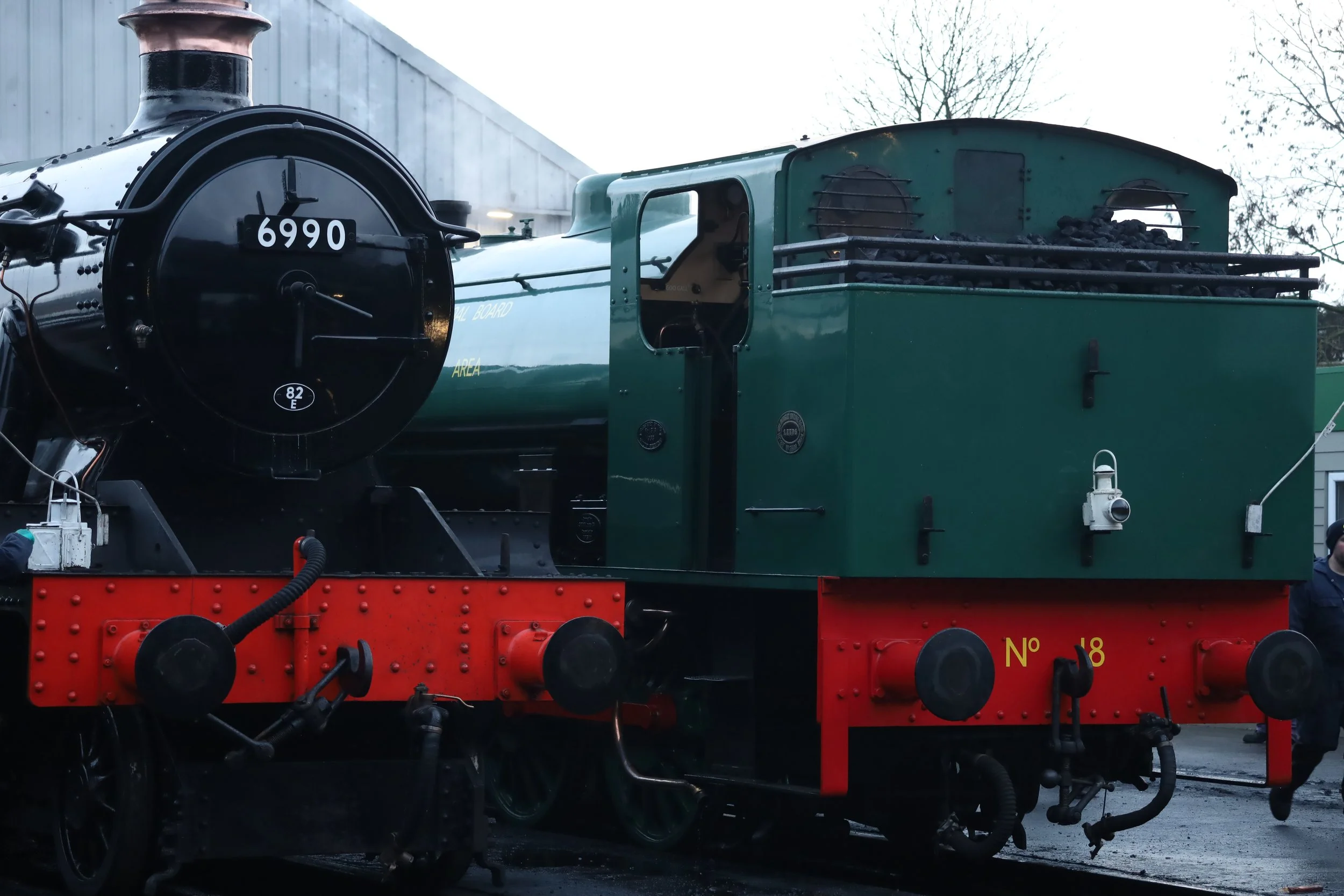 A vintage black and green steam locomotive with the number 6990 on the front, mounted on train tracks outdoors, with trees and a building in the background.